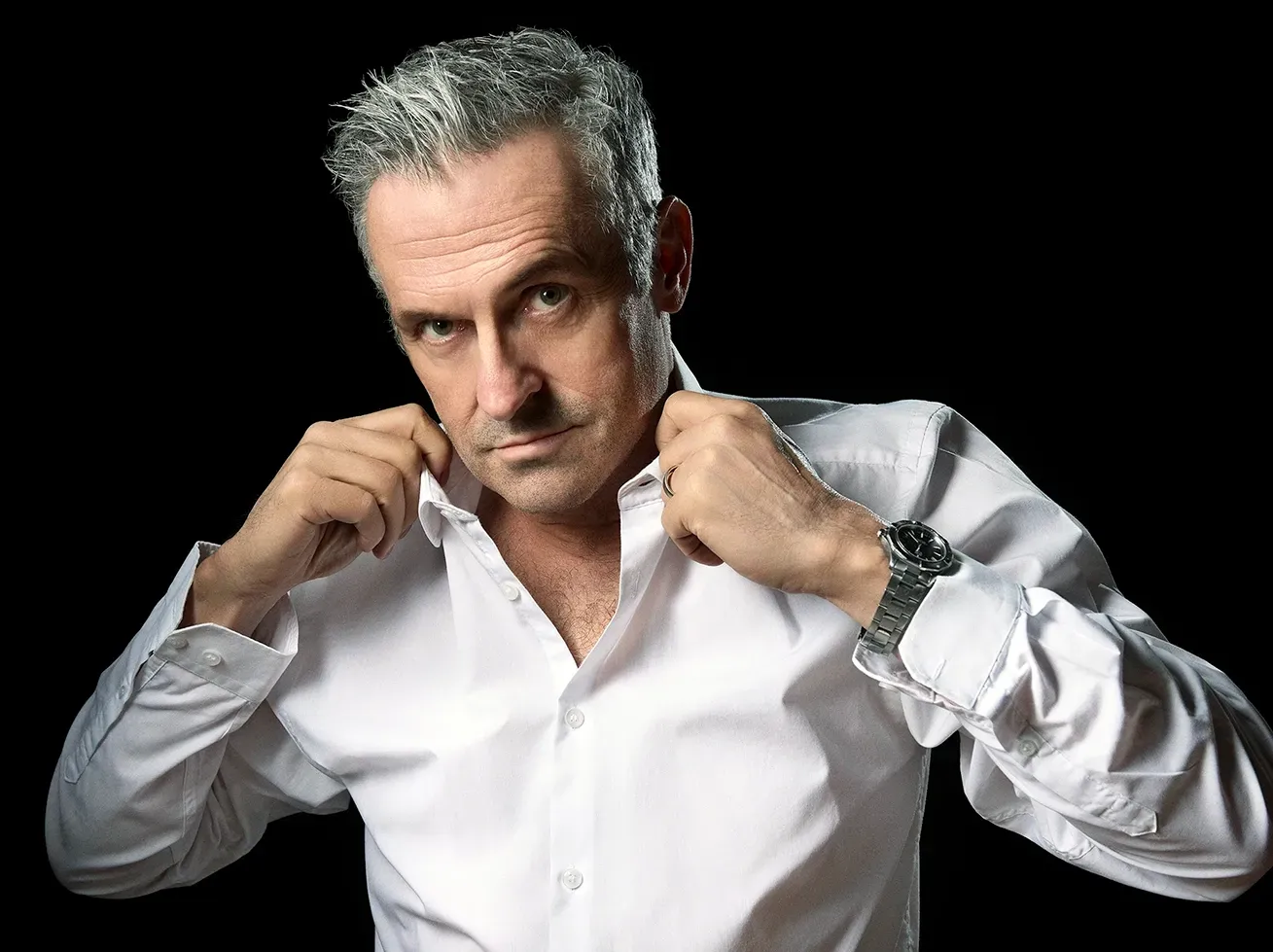 Man with gray hair and white shirt, adjusting his collar, dark background.