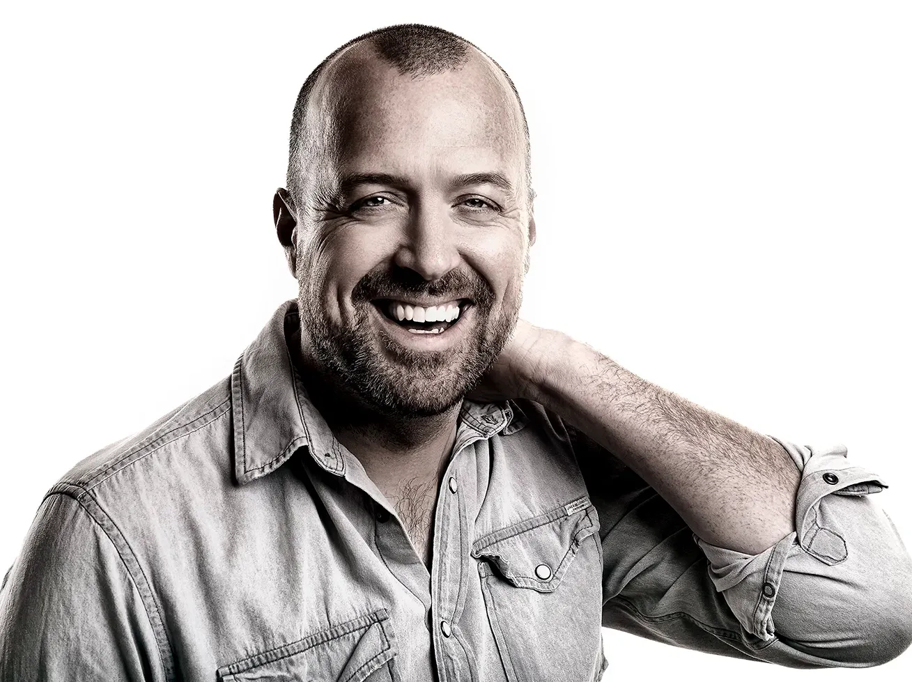 Man with a bald head and beard smiles while wearing a denim shirt, hand behind his neck.