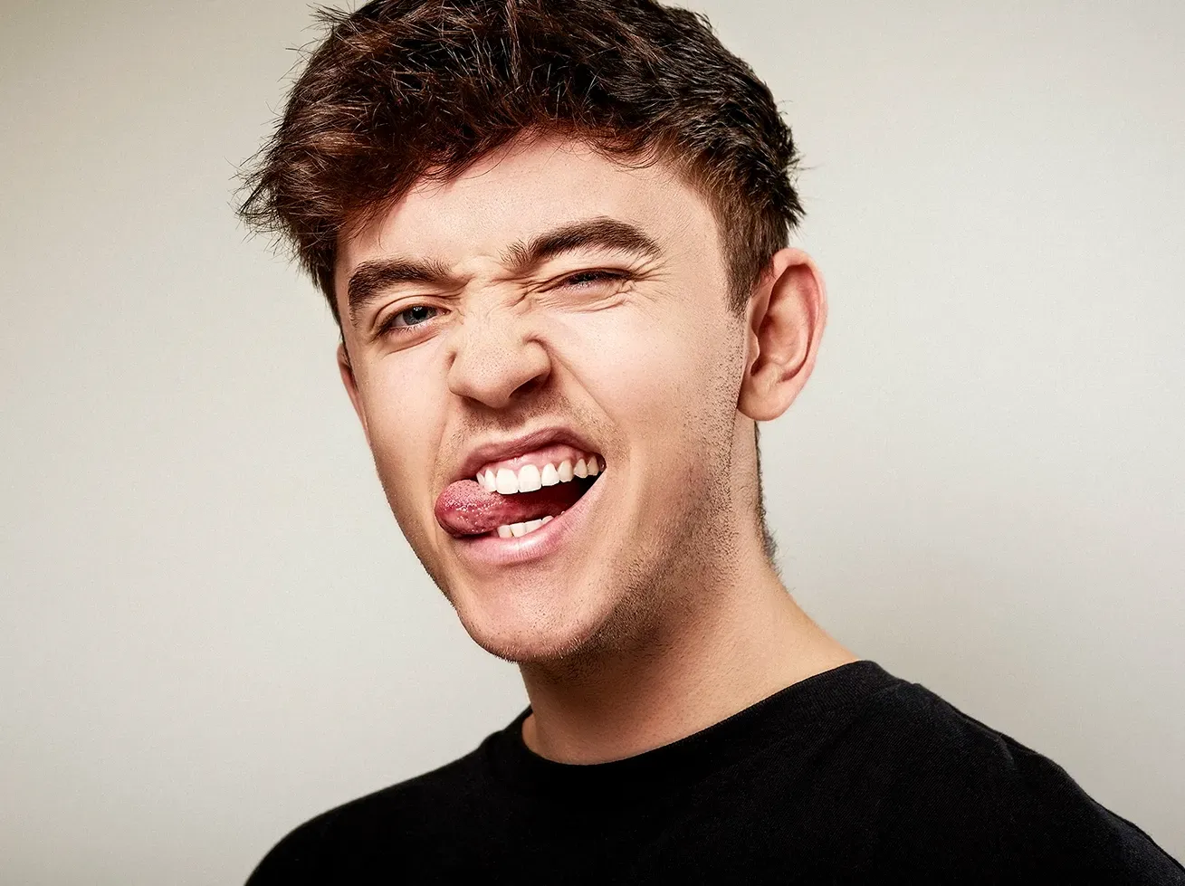 Man with dark hair winking and sticking out his tongue against a light gray background.