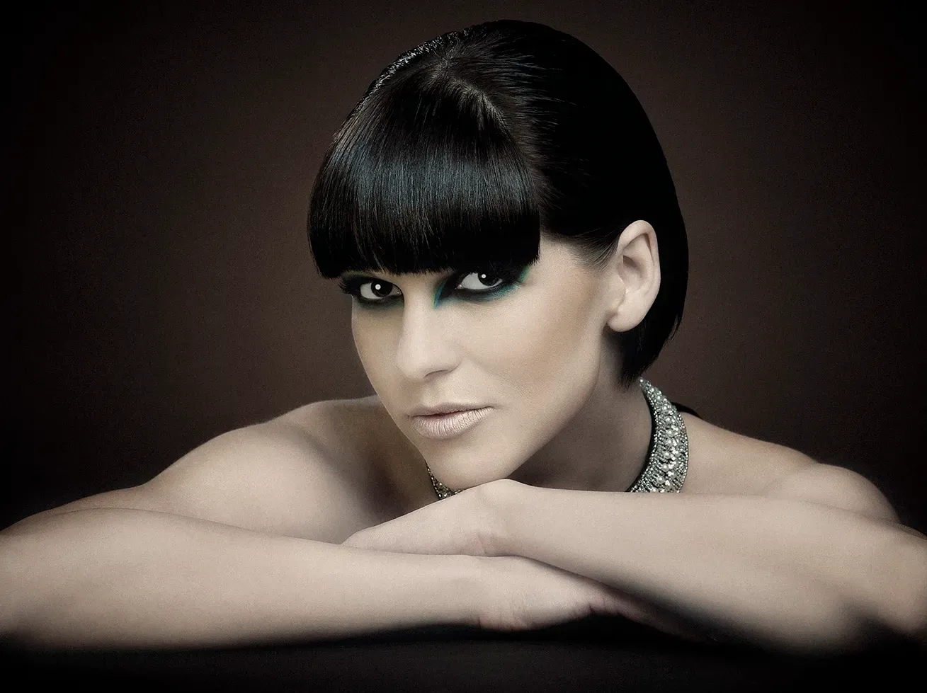 Woman with dark hair and bangs, resting arms, wearing jewelry; moody studio shot.