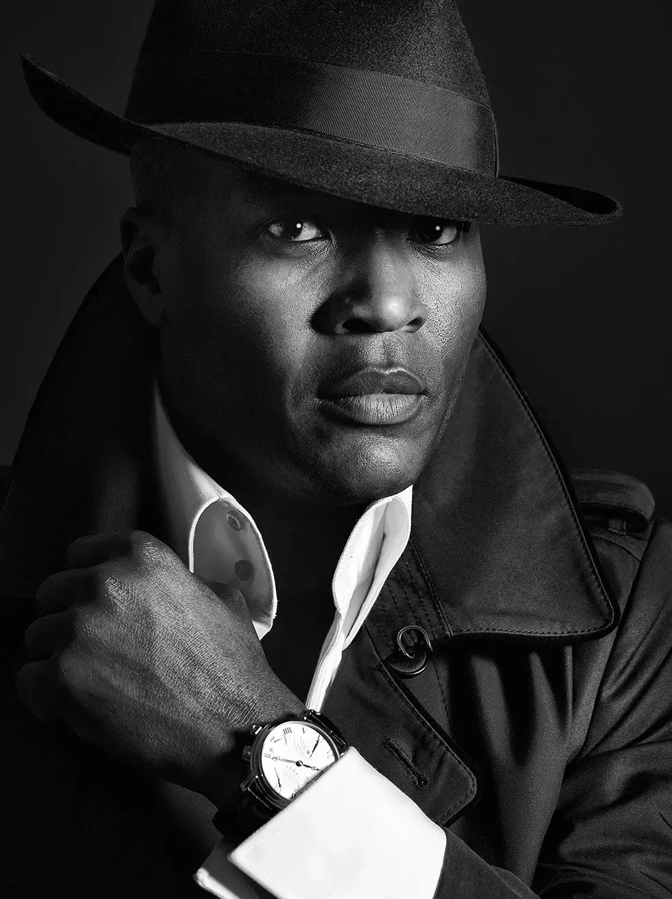 Man in fedora and coat, peering intently, wearing a watch. Black and white.