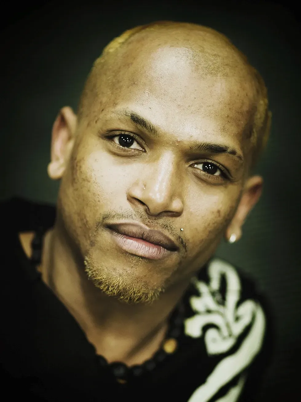 Bald man with short, light-colored facial hair, looking at the viewer. He wears a dark shirt with white detailing.