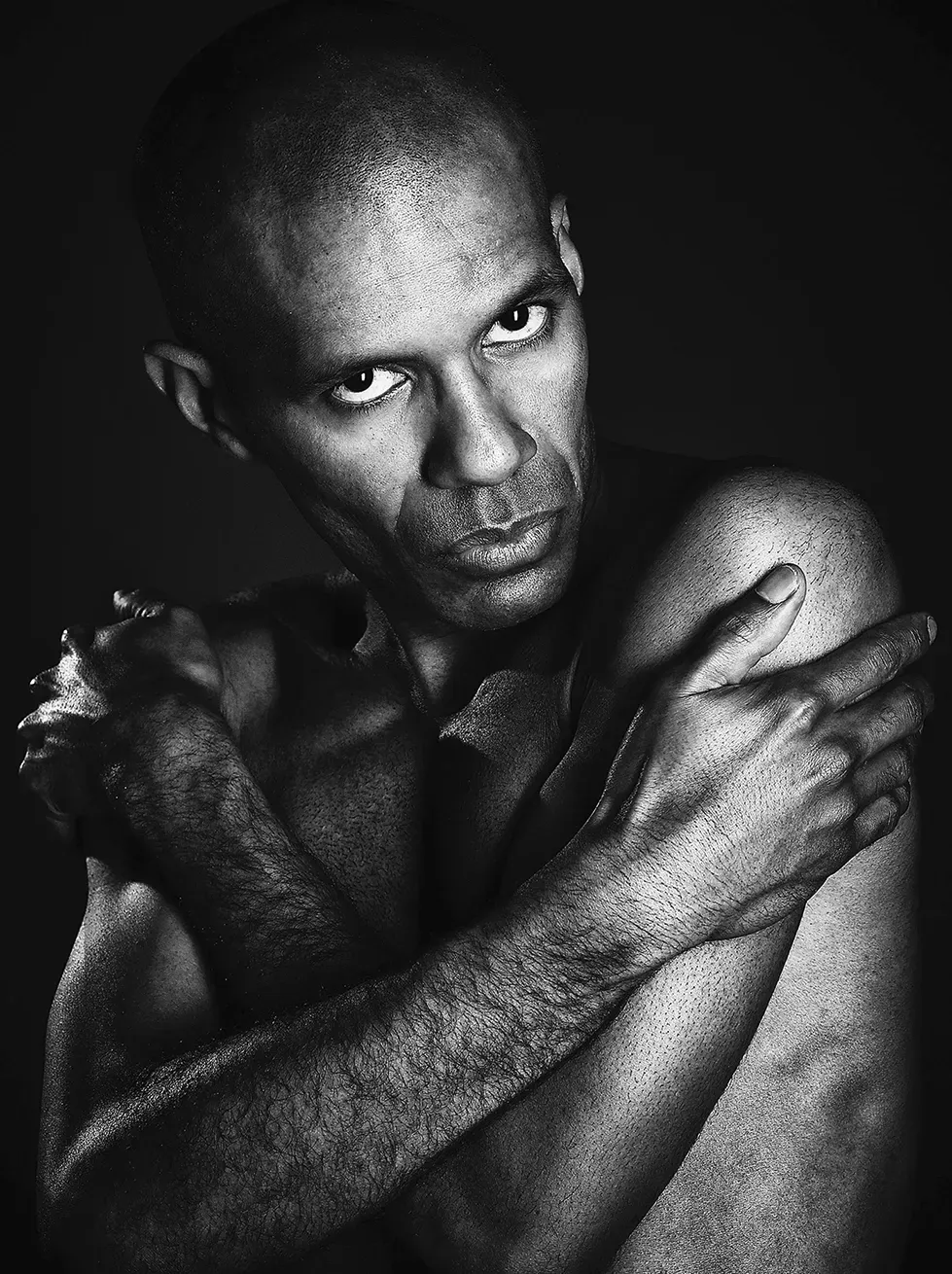 Bald person, torso visible, arms crossed around chest, looking intently at the viewer against a dark background.
