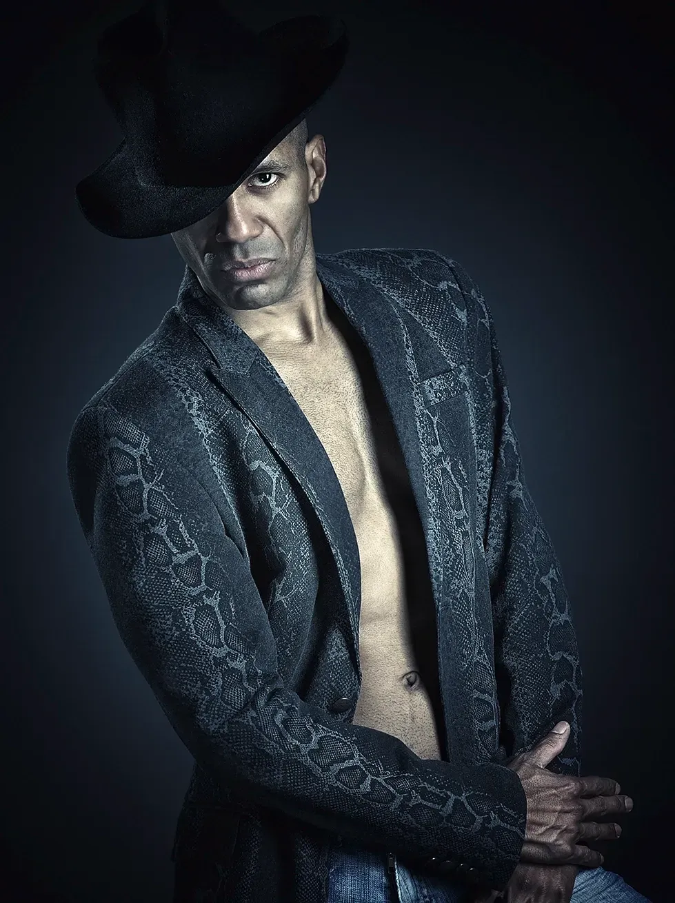 Man wearing a patterned jacket, a hat, and jeans, striking a pose.