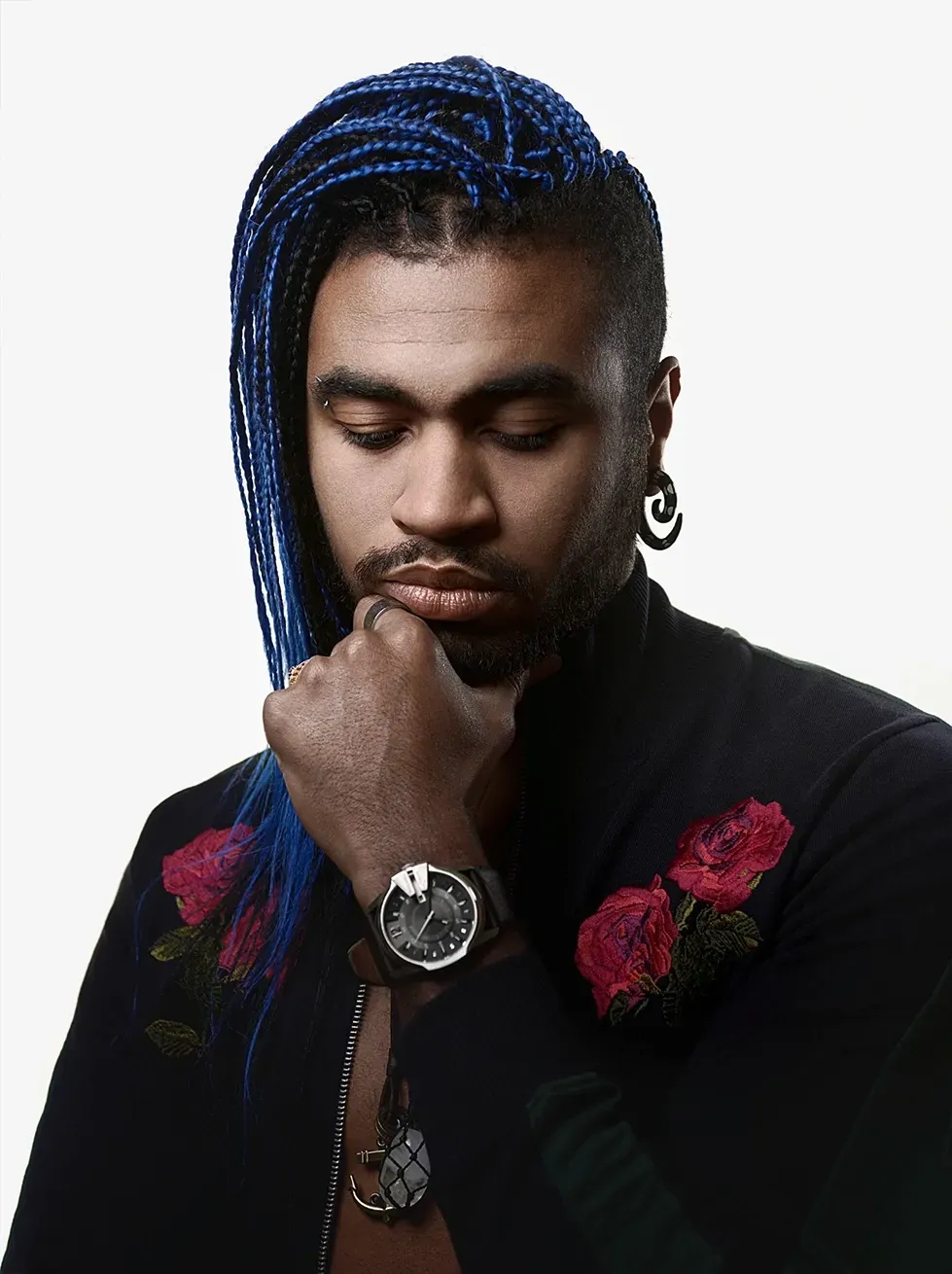 Man with blue braided hair, holding chin, wearing black jacket with red roses, large earrings, and watch.