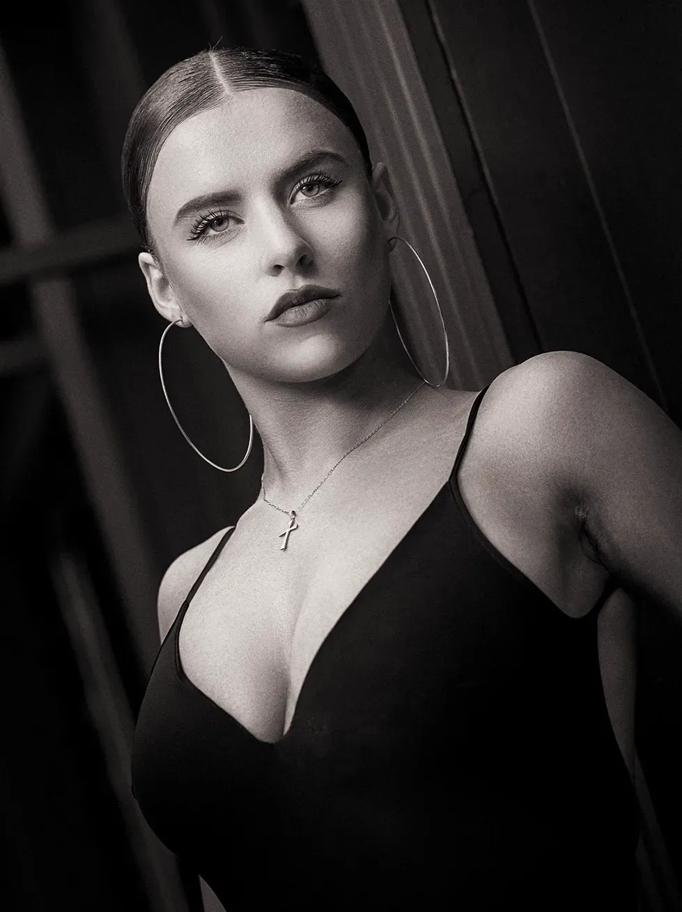 Woman in a black dress, large hoop earrings, and necklace, looking upwards with a serious expression.