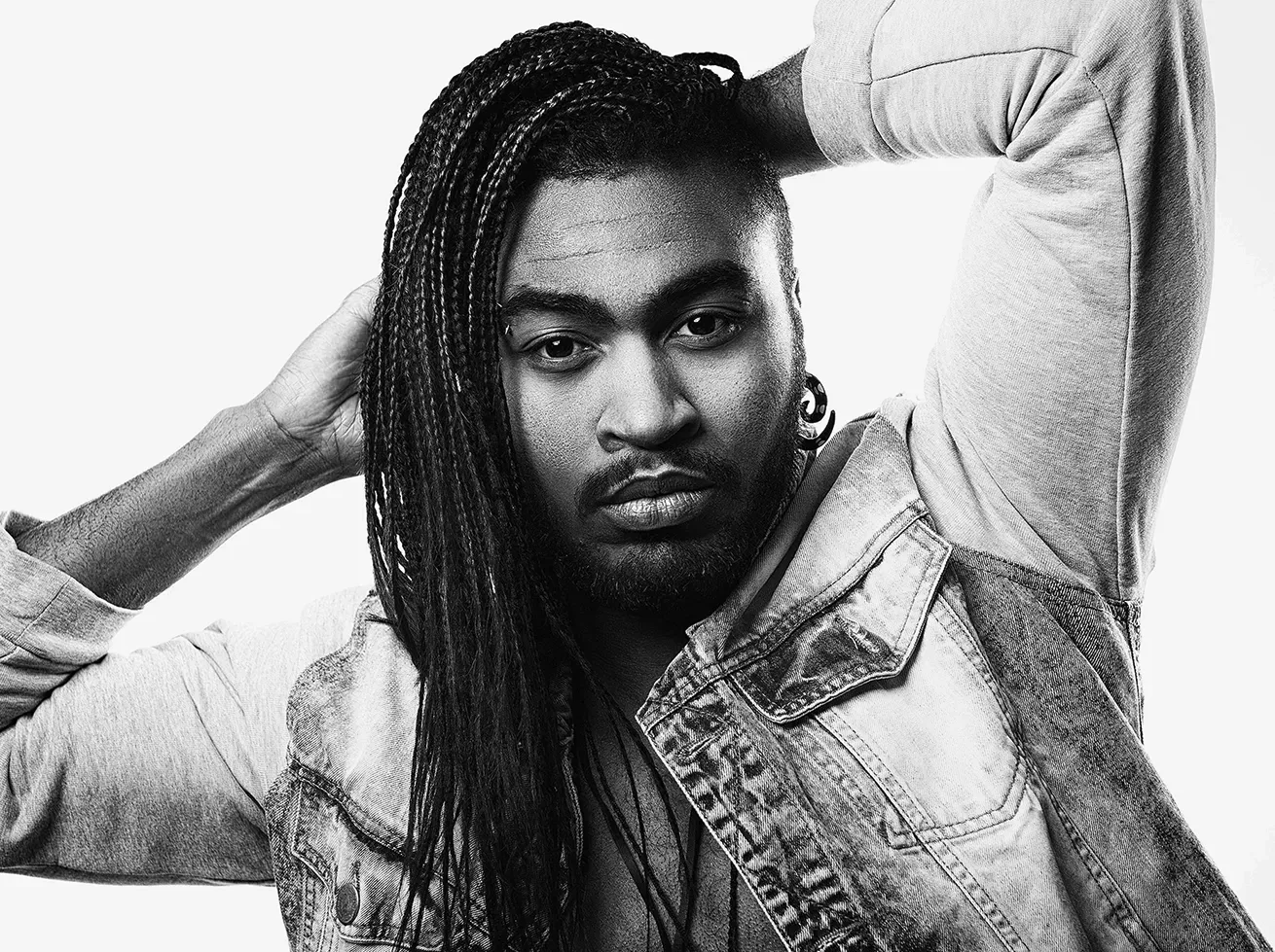 Man with long, braided hair, wearing a denim jacket, arms raised behind head, staring forward.