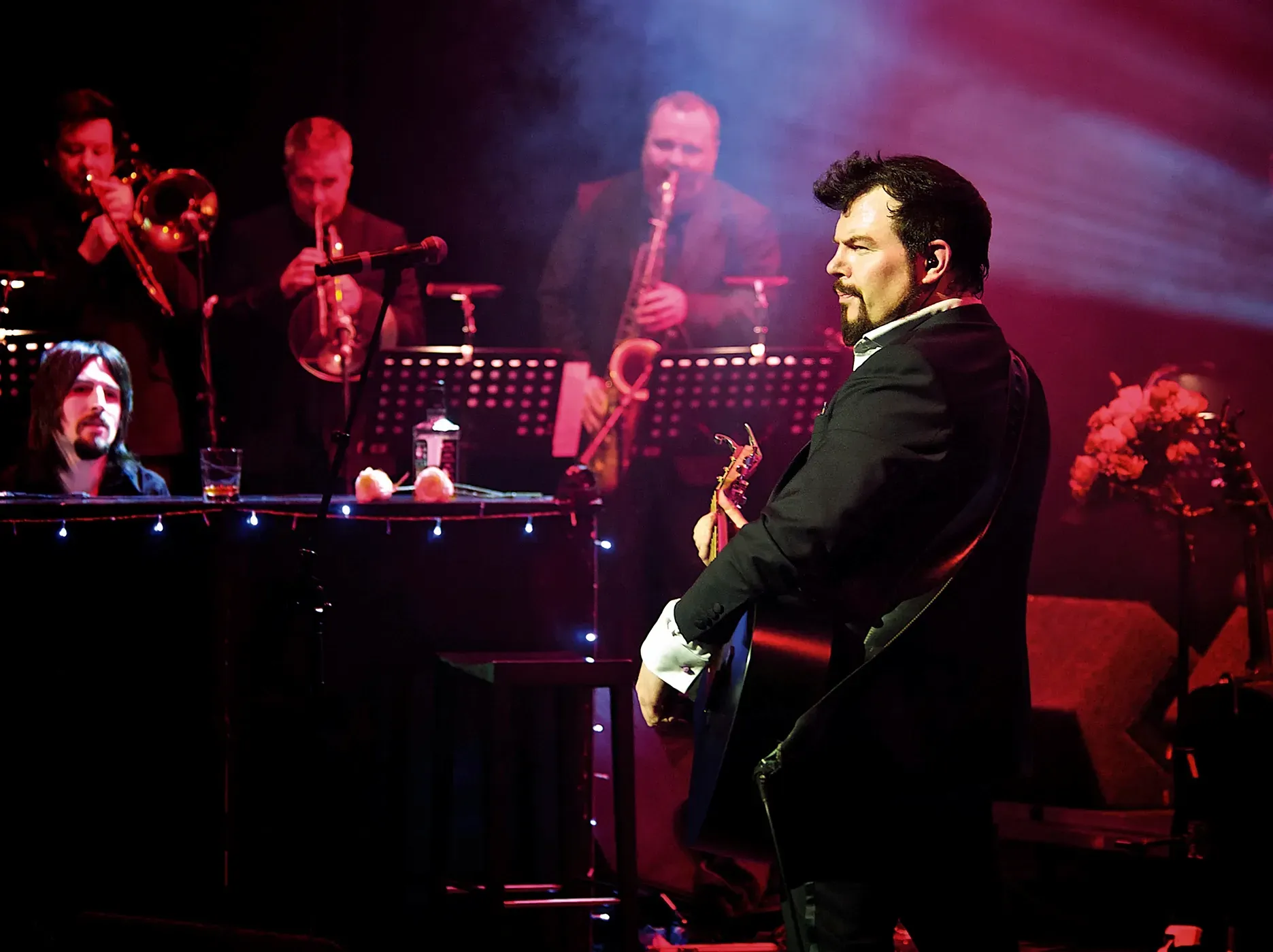 Band performing on stage: Musician in suit plays guitar, band members with brass instruments in background.