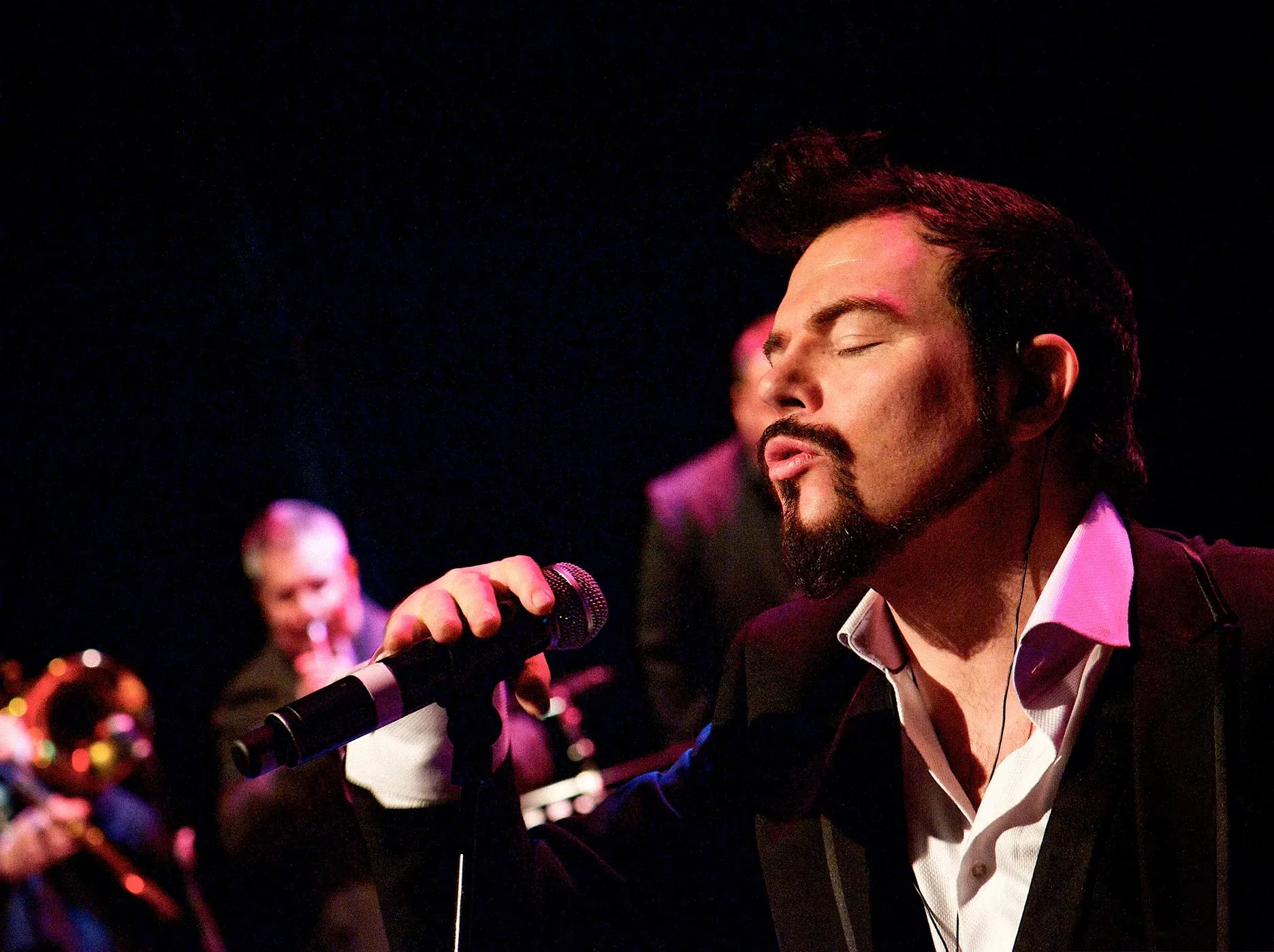 A man with a goatee sings into a microphone on stage, eyes closed, with a band in the background.