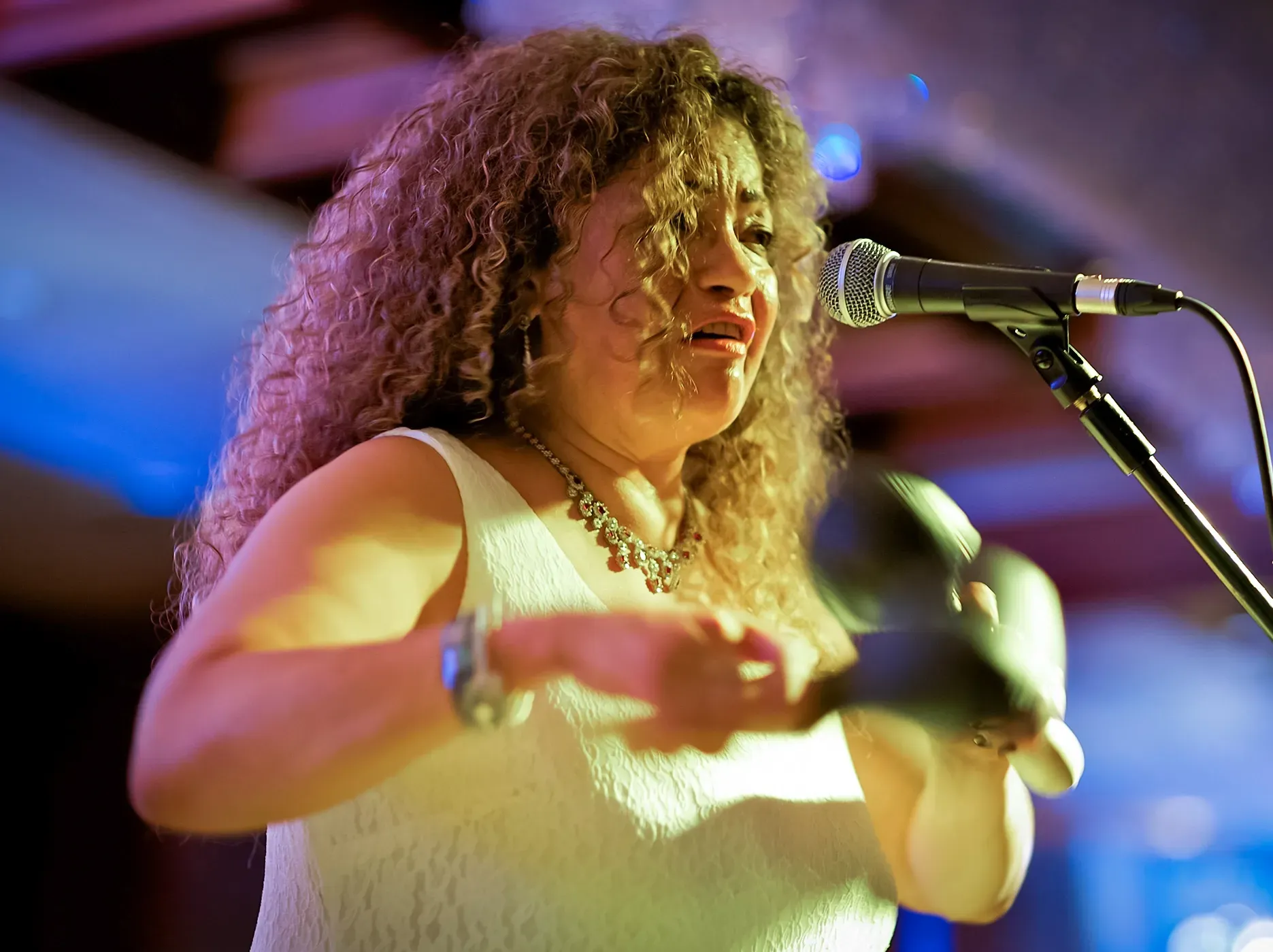 Woman singing into a microphone, playing maracas, wearing a white dress and silver jewelry.