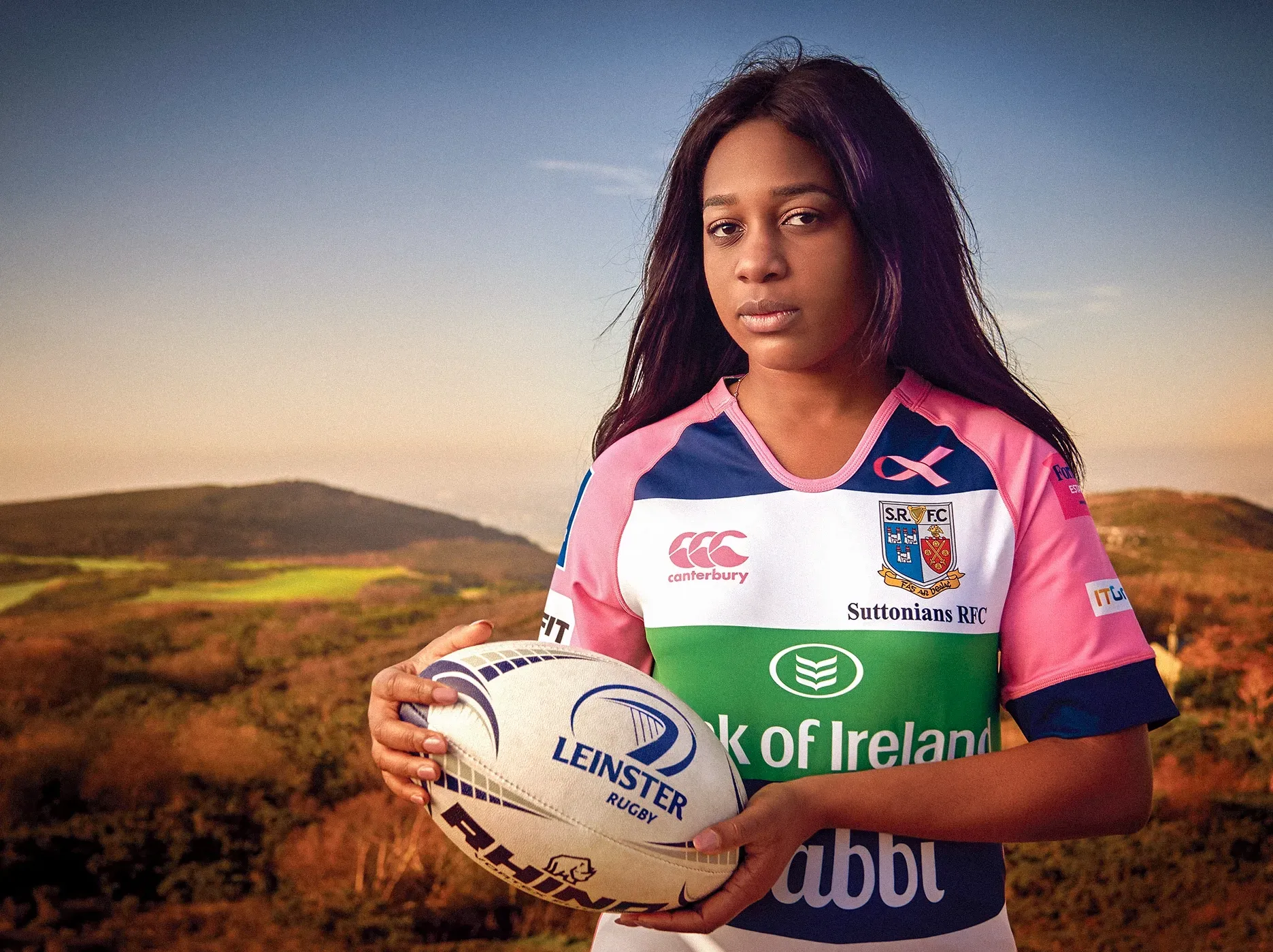 Woman holding a rugby ball, wearing a pink and white jersey with a green stripe, against a scenic background.