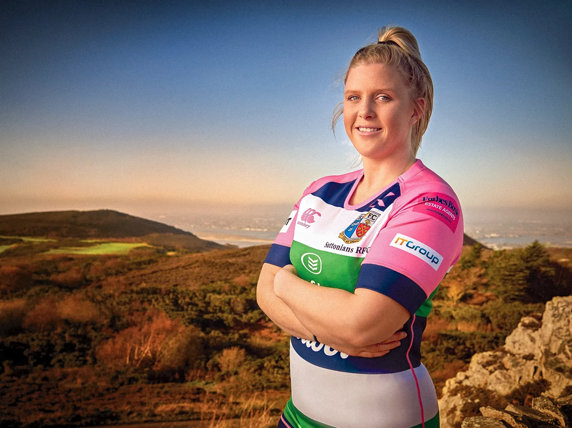 Woman in rugby jersey stands on a hilltop, arms crossed, smiling. Landscape background with mountains.