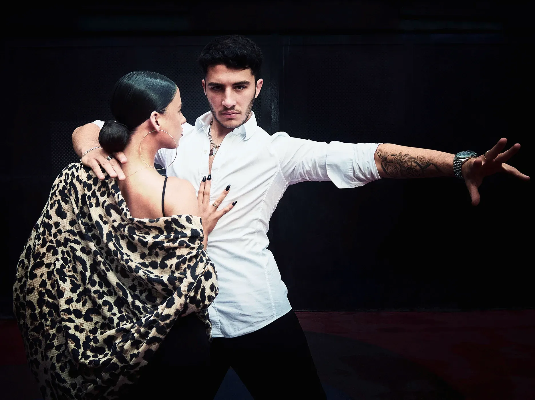 Couple dancing close together, man in white shirt, arm outstretched; woman in leopard print. Black background.