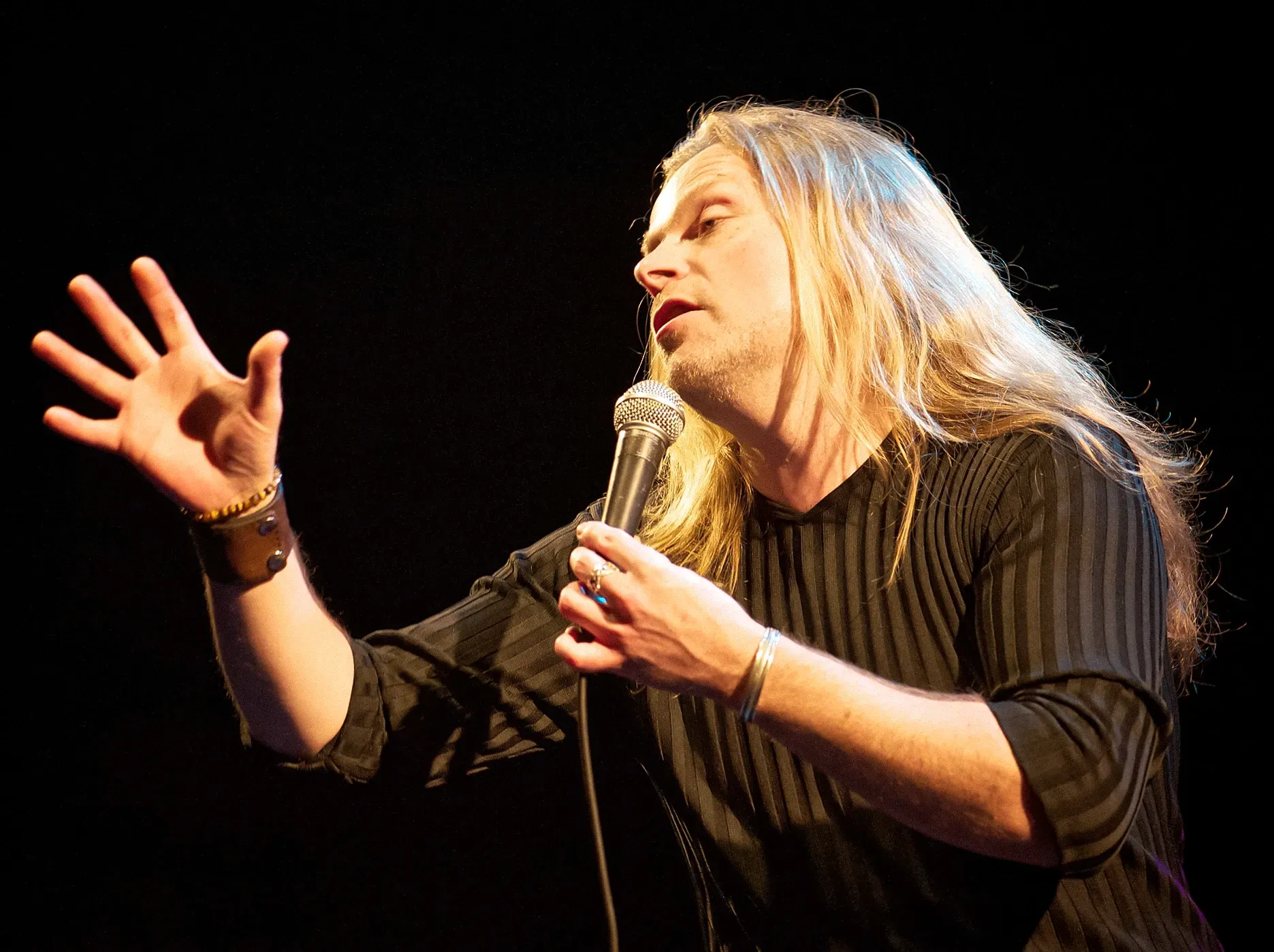 Man with long blonde hair, holding a microphone, gesturing with his hand. Black shirt, dark background.