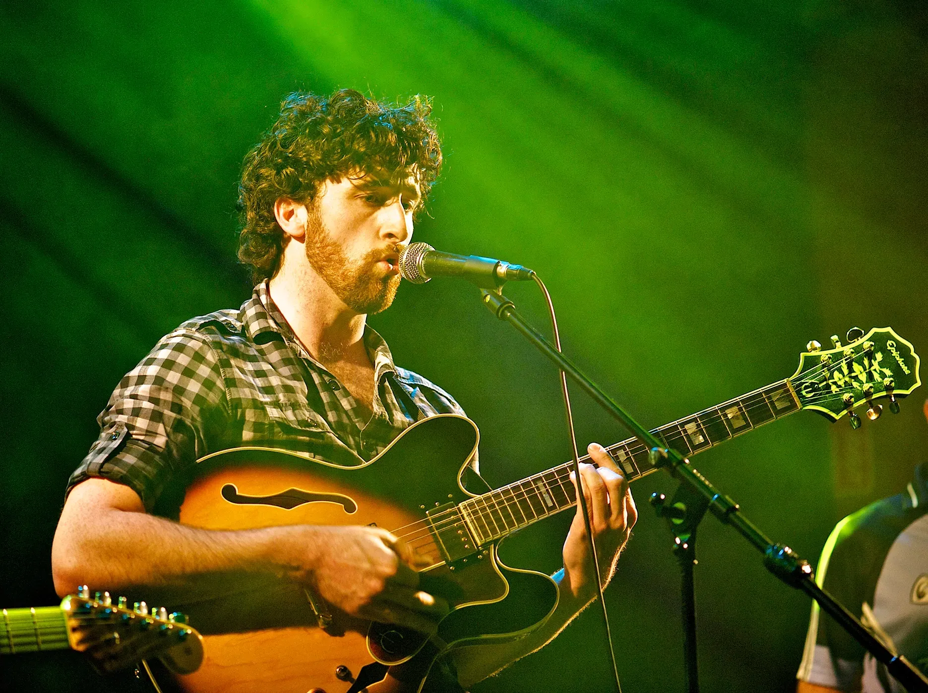 Guitarist singing into a microphone onstage under green lighting. Wearing a plaid shirt, holding a hollow-body guitar.