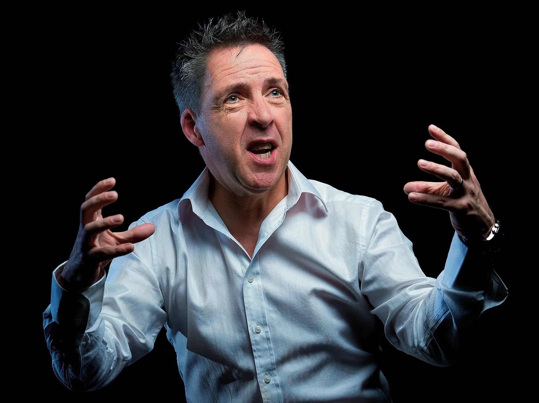 Man in white shirt with open mouth and raised hands, against a black background.