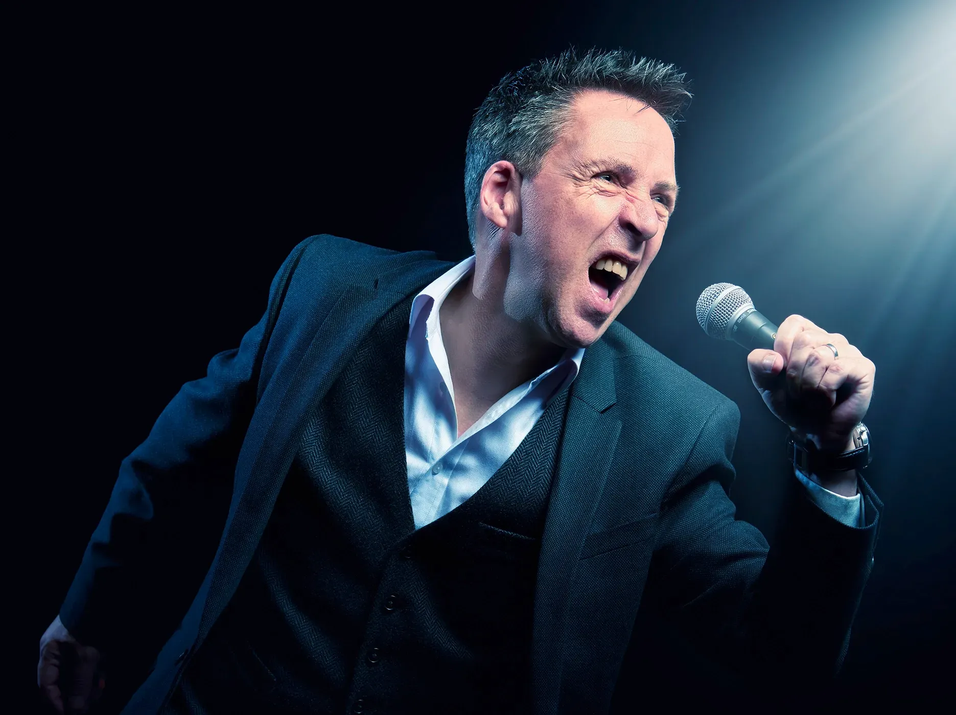 Man singing into a microphone, lit by a spotlight, wearing a suit, against a dark background.