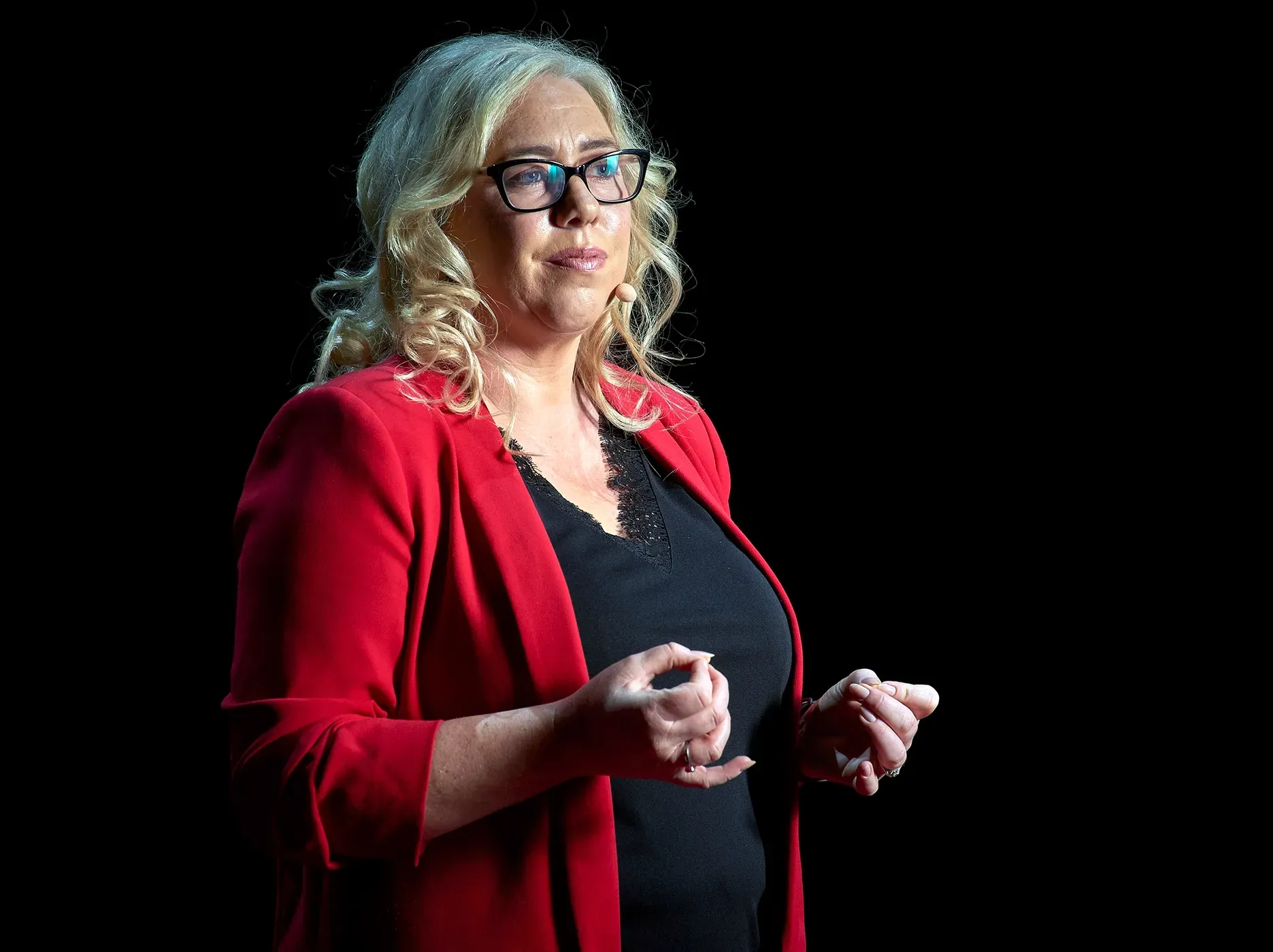 Woman in red jacket and glasses speaking against a black background.