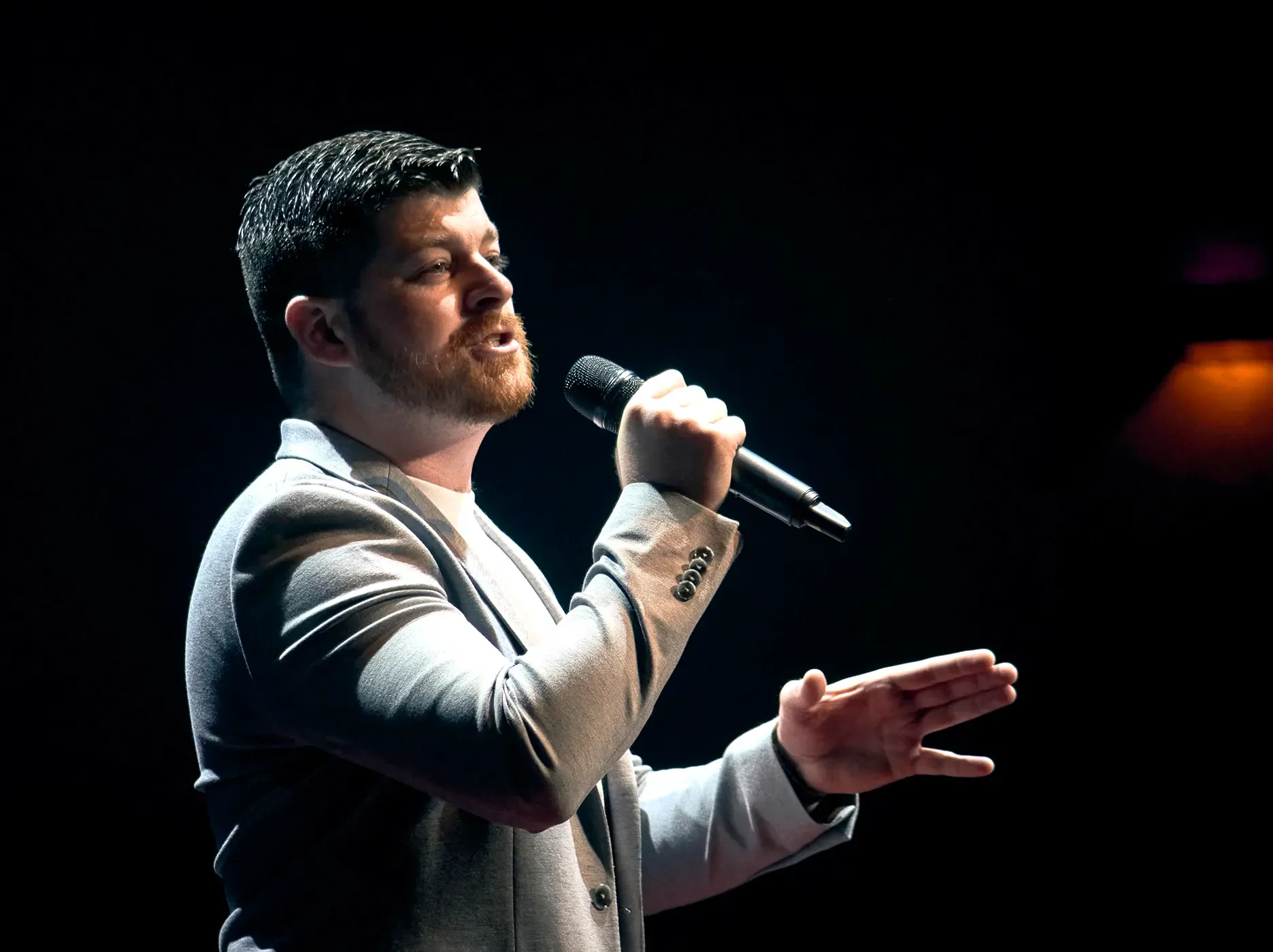 Man singing into a microphone on stage, wearing a gray suit jacket, gesturing with his hand.