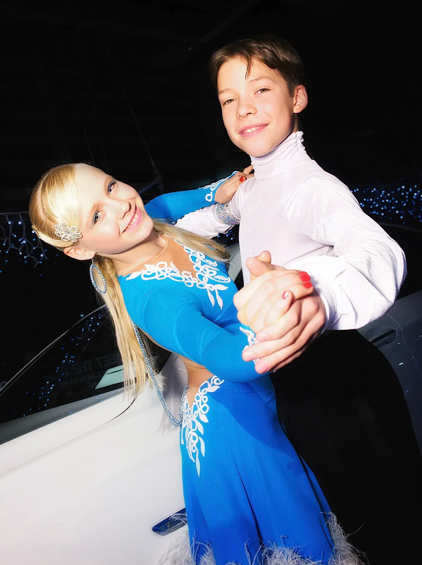 Young couple in formal wear dancing, girl in blue dress, boy in white shirt, smiling, lit background.