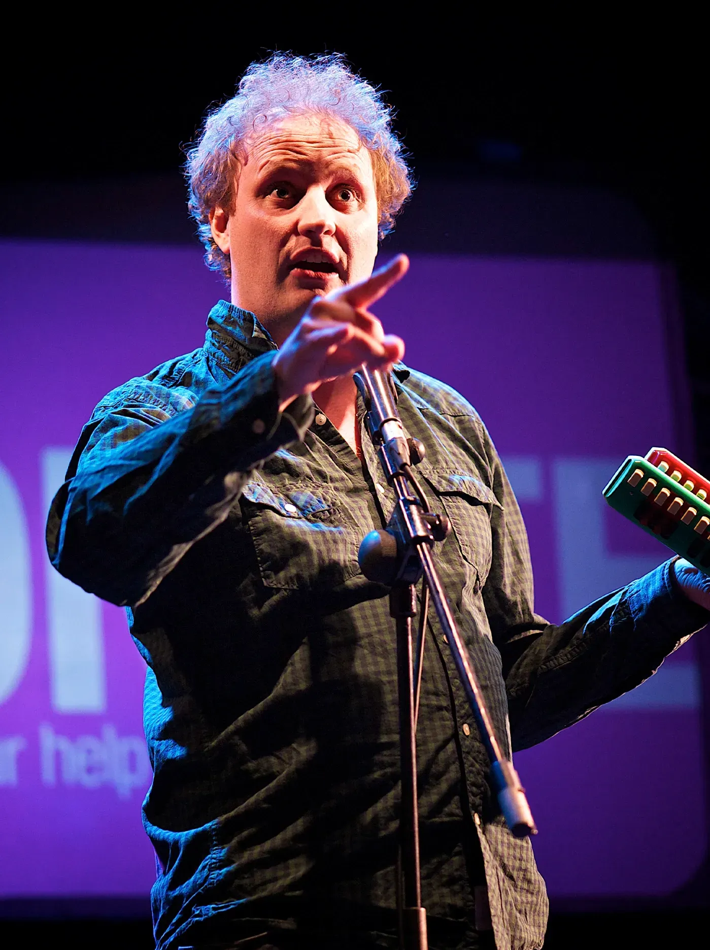 Man on stage with a microphone, pointing, holding a hand percussion instrument, purple backdrop.