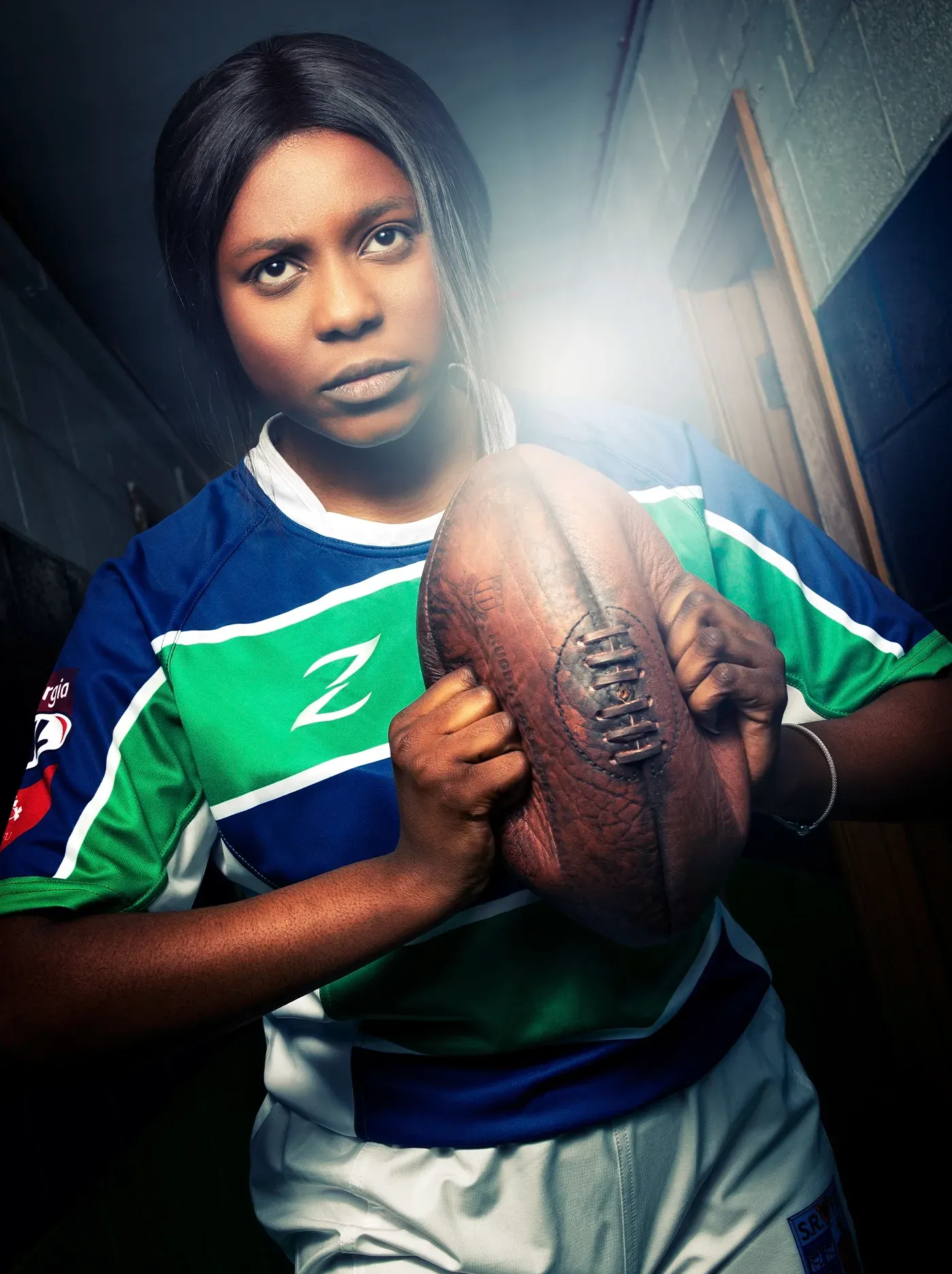 Woman in sports jersey holding a football, lit by bright light in a hallway.