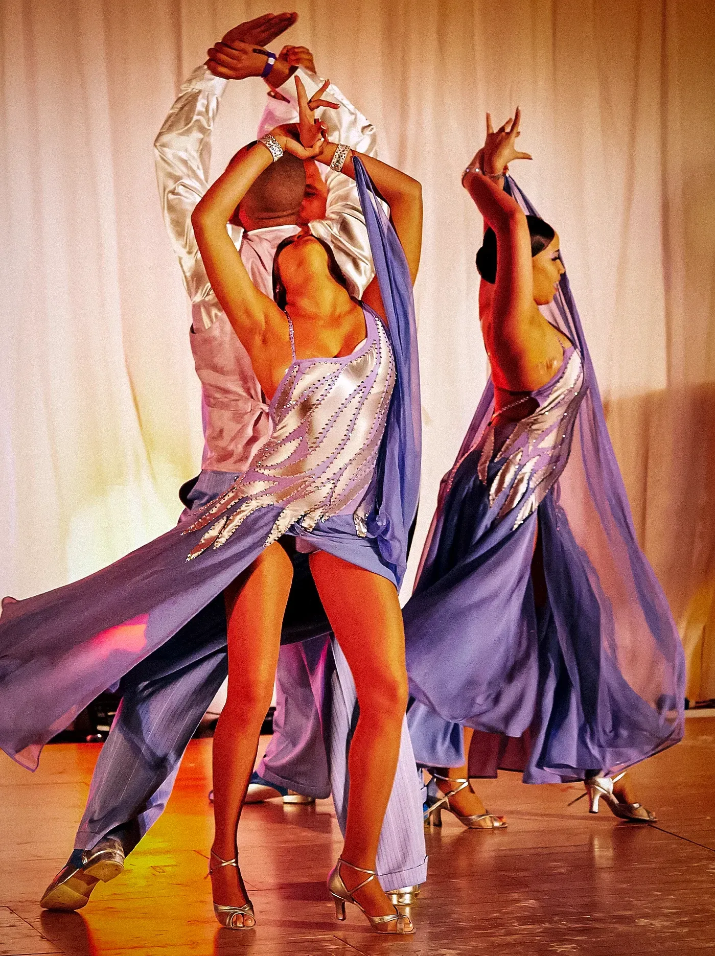 Dancers in blue and silver gowns performing on stage, arms raised, in front of a white curtain.