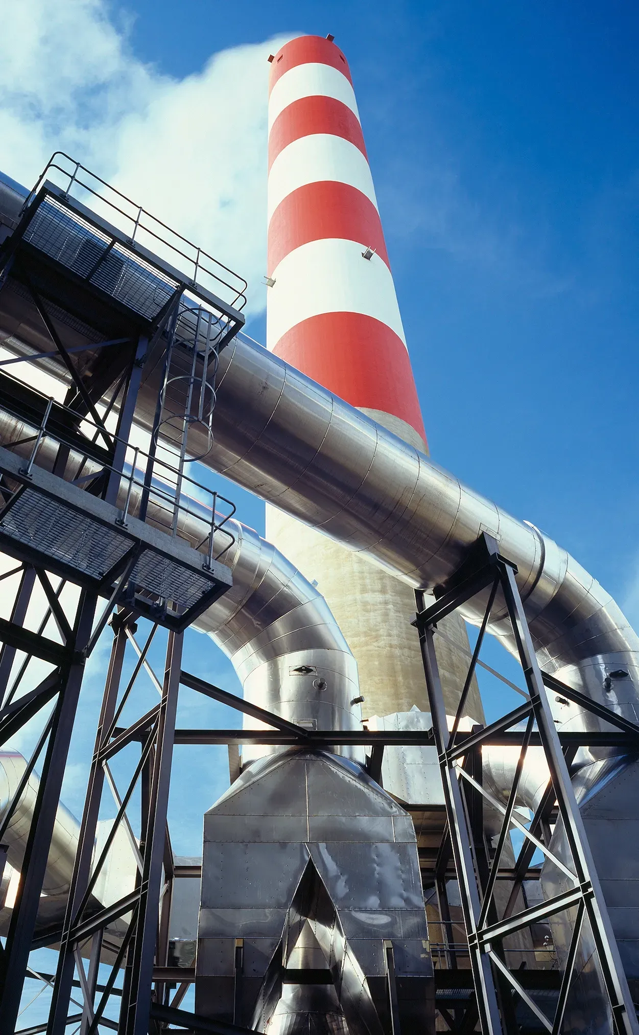 Red and white striped smokestack with silver pipes and supports, against a blue sky.