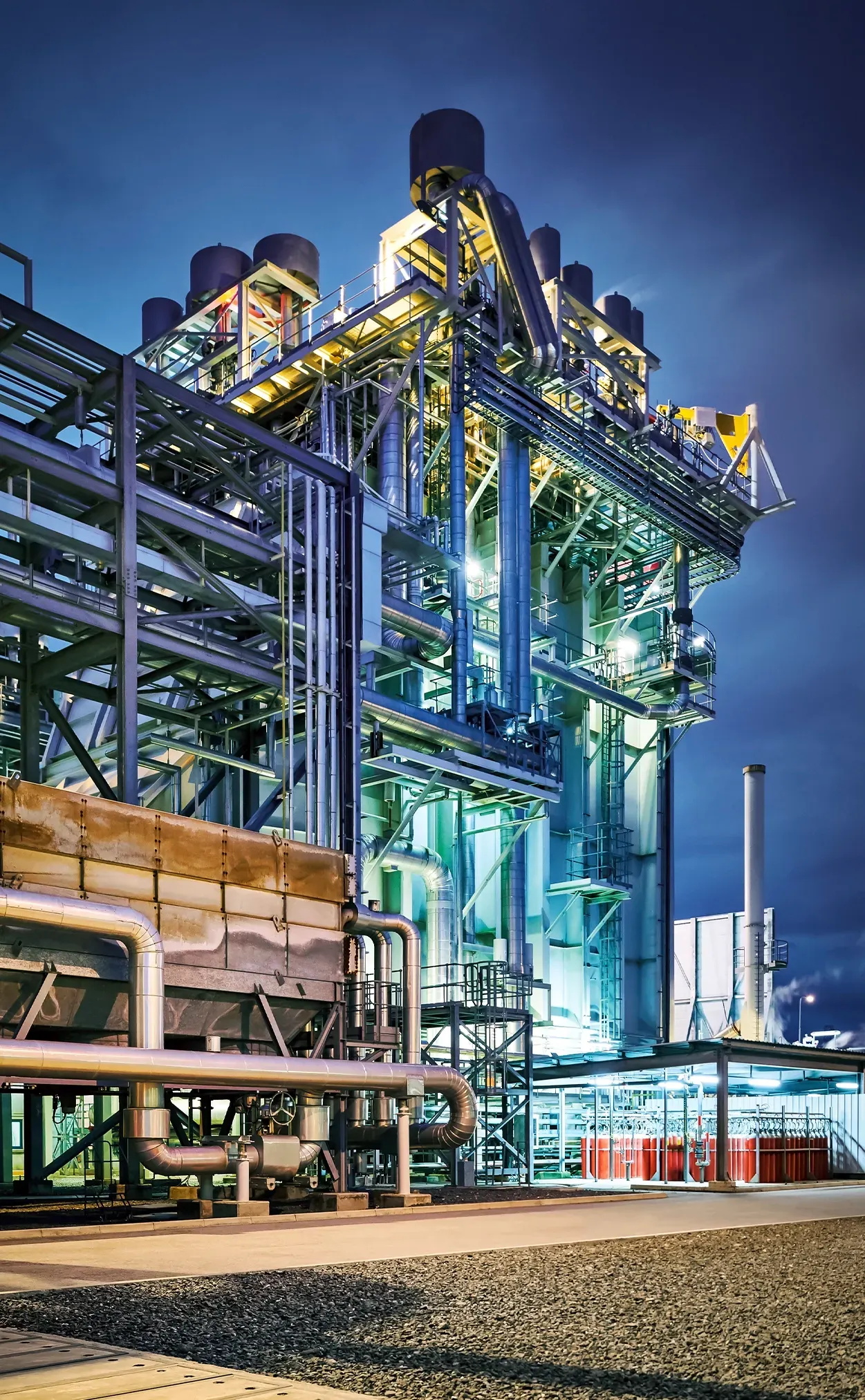 Power plant at dusk, illuminated by bright lights, complex structure of pipes and machinery.