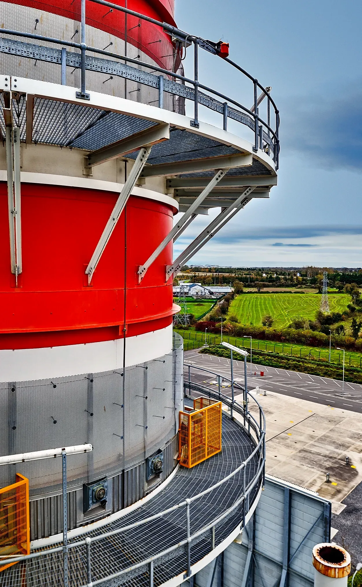 Close-up of a red and white industrial tower with a walkway and a view of green fields and a road.