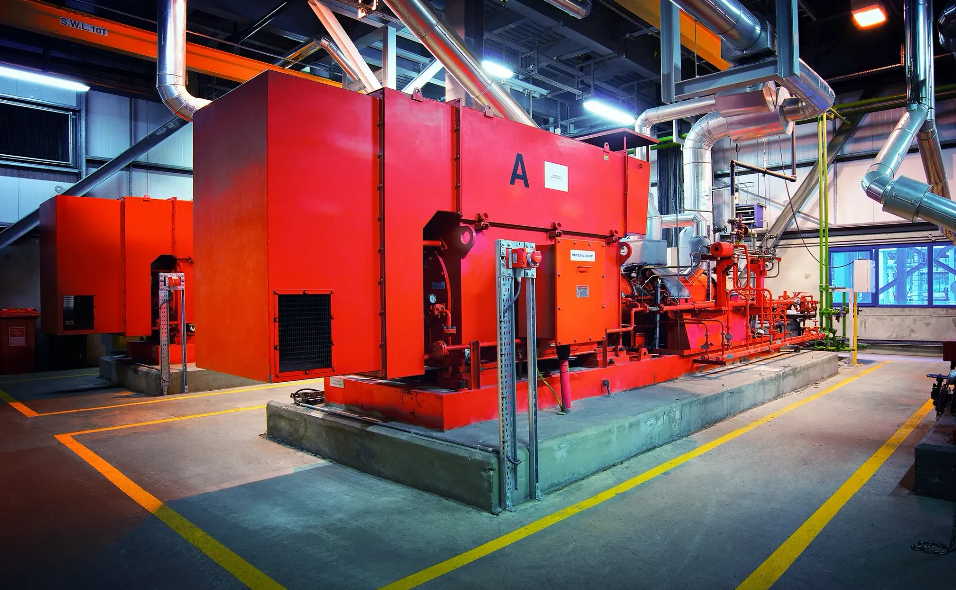 Red industrial machinery in a factory setting. Pipes and ventilation visible. Yellow safety lines on floor.
