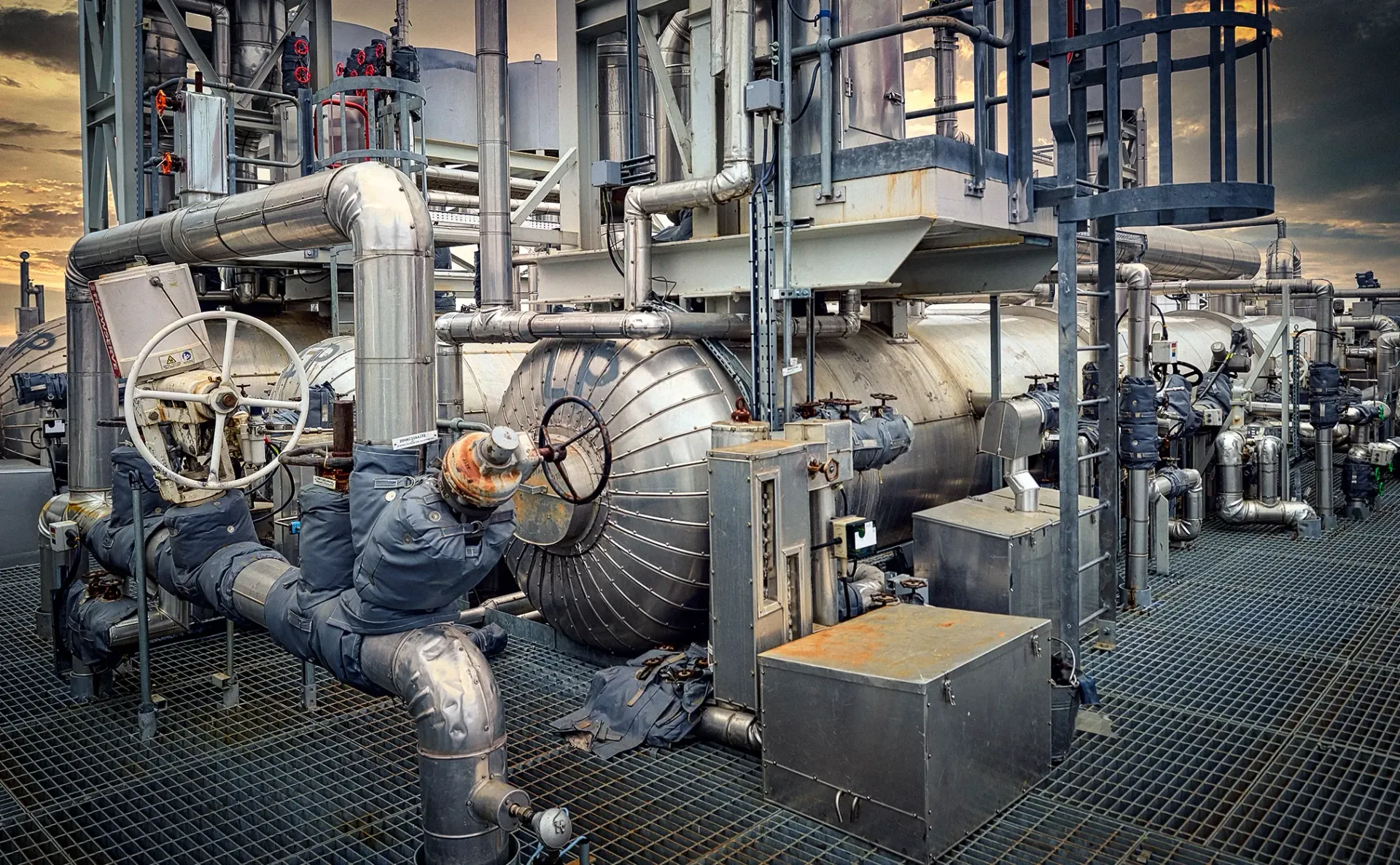 Industrial complex with metal pipes, tanks, and platform; cloudy sky.
