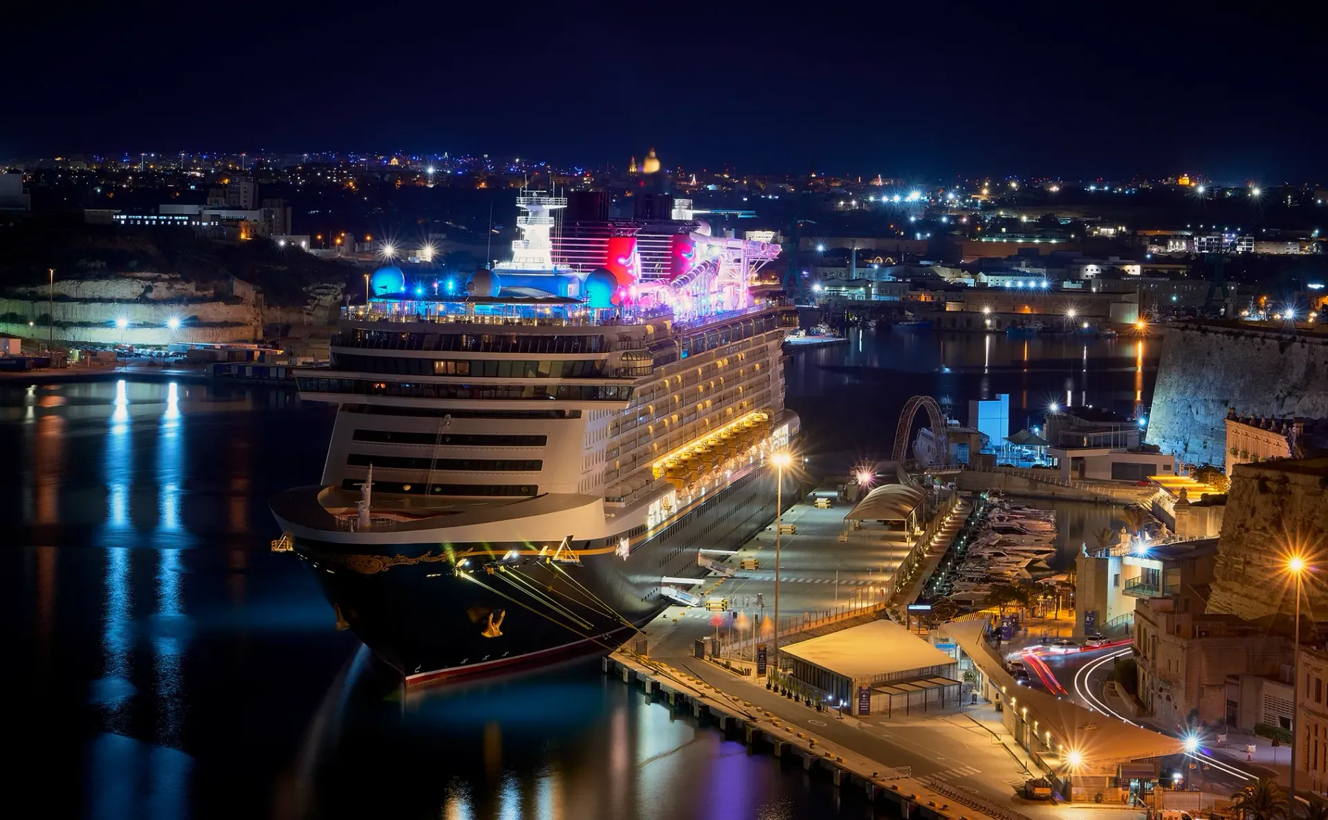Cruise ship docked at a city harbor at night with lights reflecting on the water.