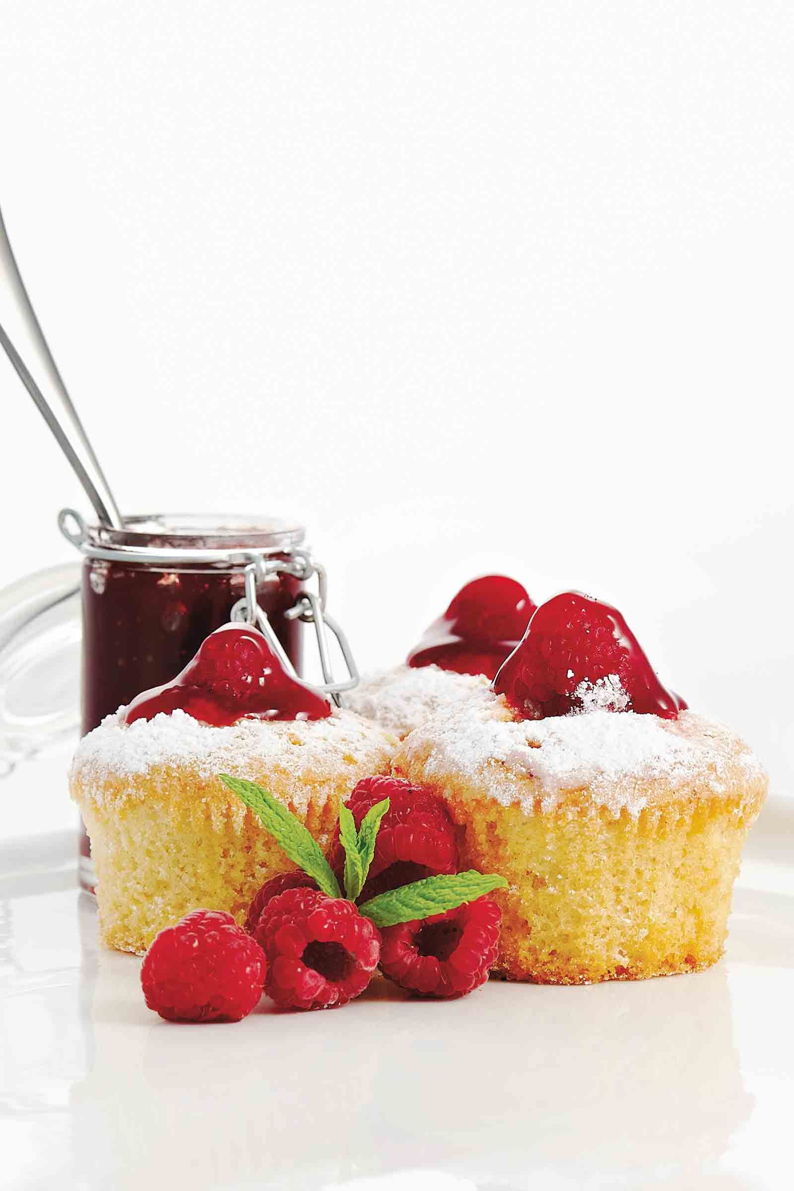 Two muffins topped with raspberries and jam, next to a jar of jam and fresh raspberries, powdered sugar, and mint.
