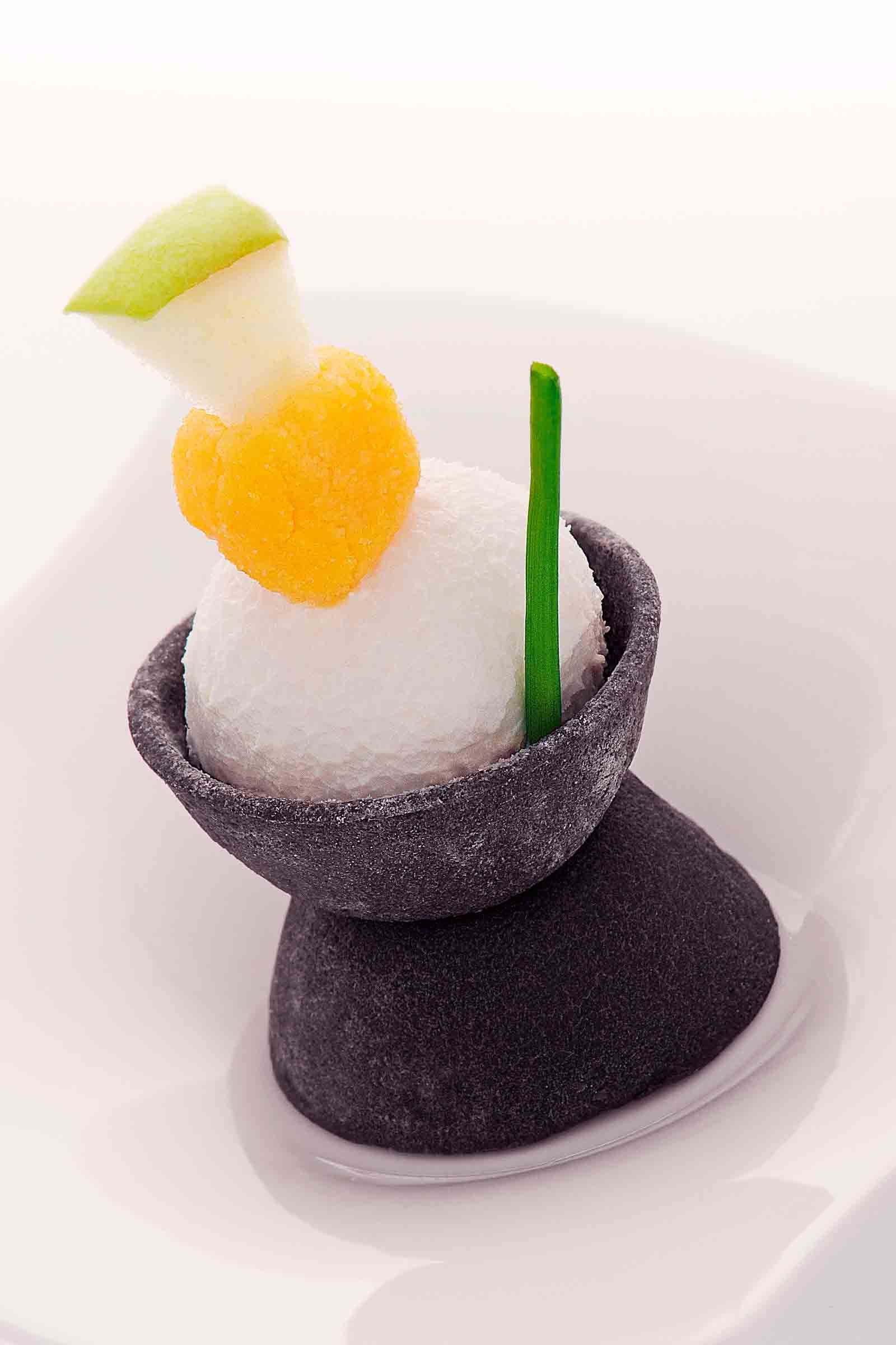 White scoop of dessert, topped with orange and green fruit, in a dark bowl on a dark pedestal, on a white plate.