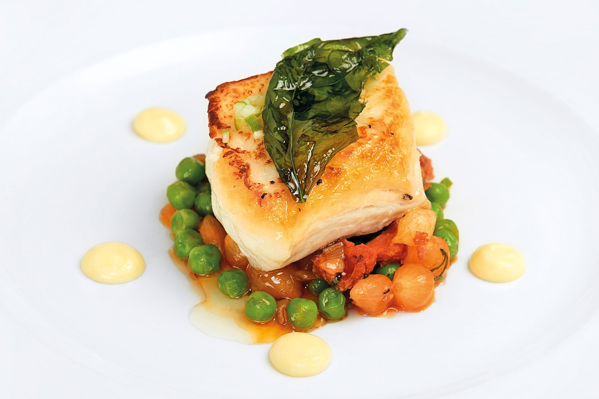 Seared fish fillet with peas and tomato, garnished with greens and sauce, on a white plate.