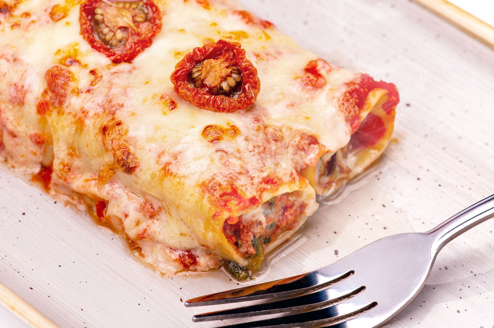 Baked cannelloni pasta with melted cheese and sundried tomatoes on a white plate with a fork.