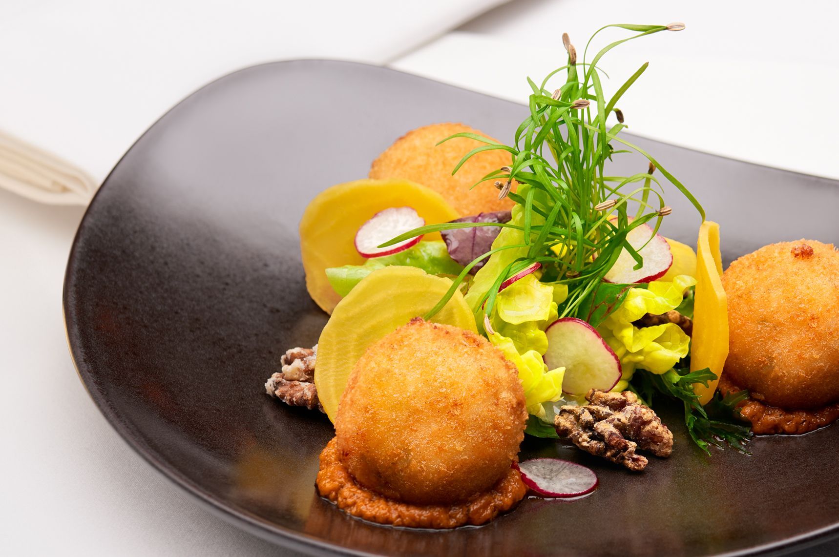 Plate of gourmet food: golden fried balls, yellow beet slices, greens, walnuts, and radish slices on a dark plate.
