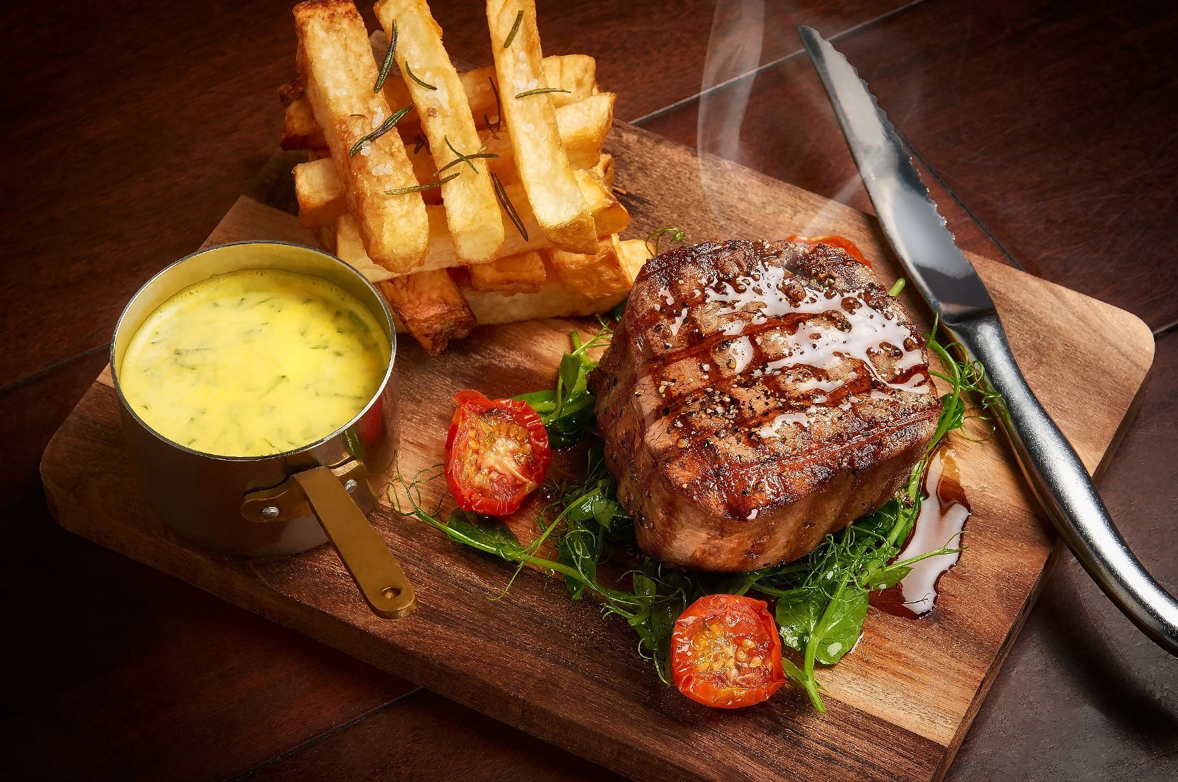 Steak with fries, sauce, and tomatoes on a wooden board with a knife.