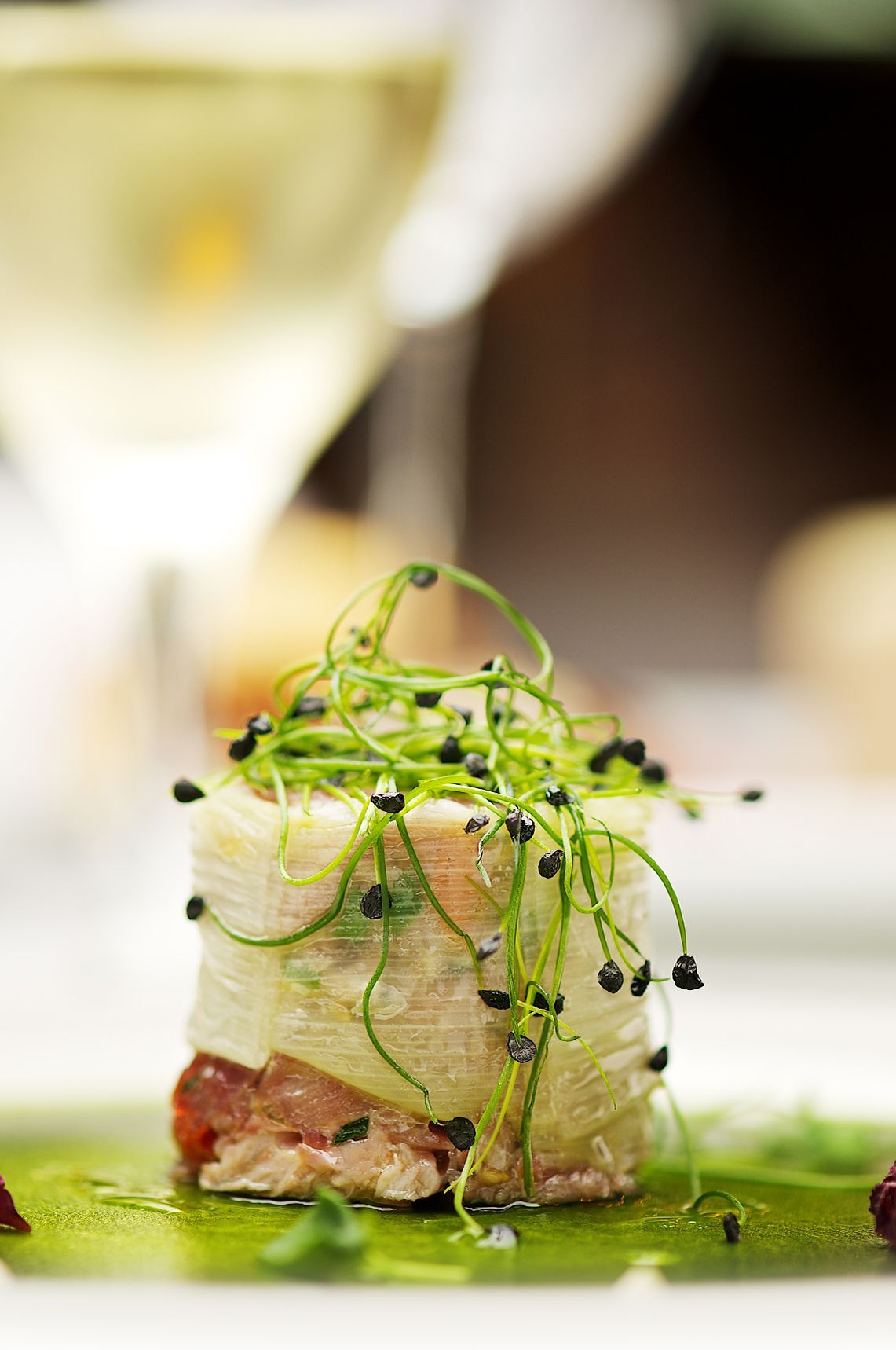 Appetizer: rolled vegetable, topped with sprouts and black seeds, over green sauce. A glass of white wine in the background.