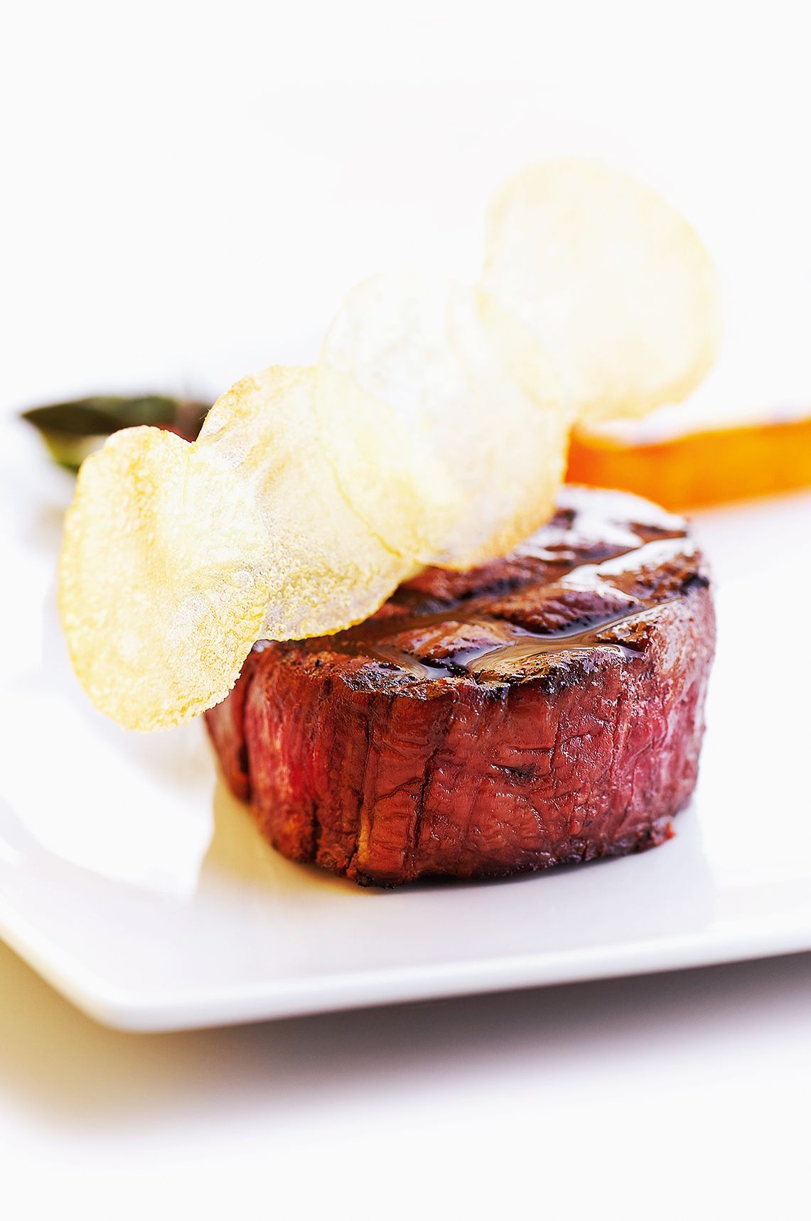 Grilled steak on a white plate, topped with a potato crisp and accompanied by side dishes.