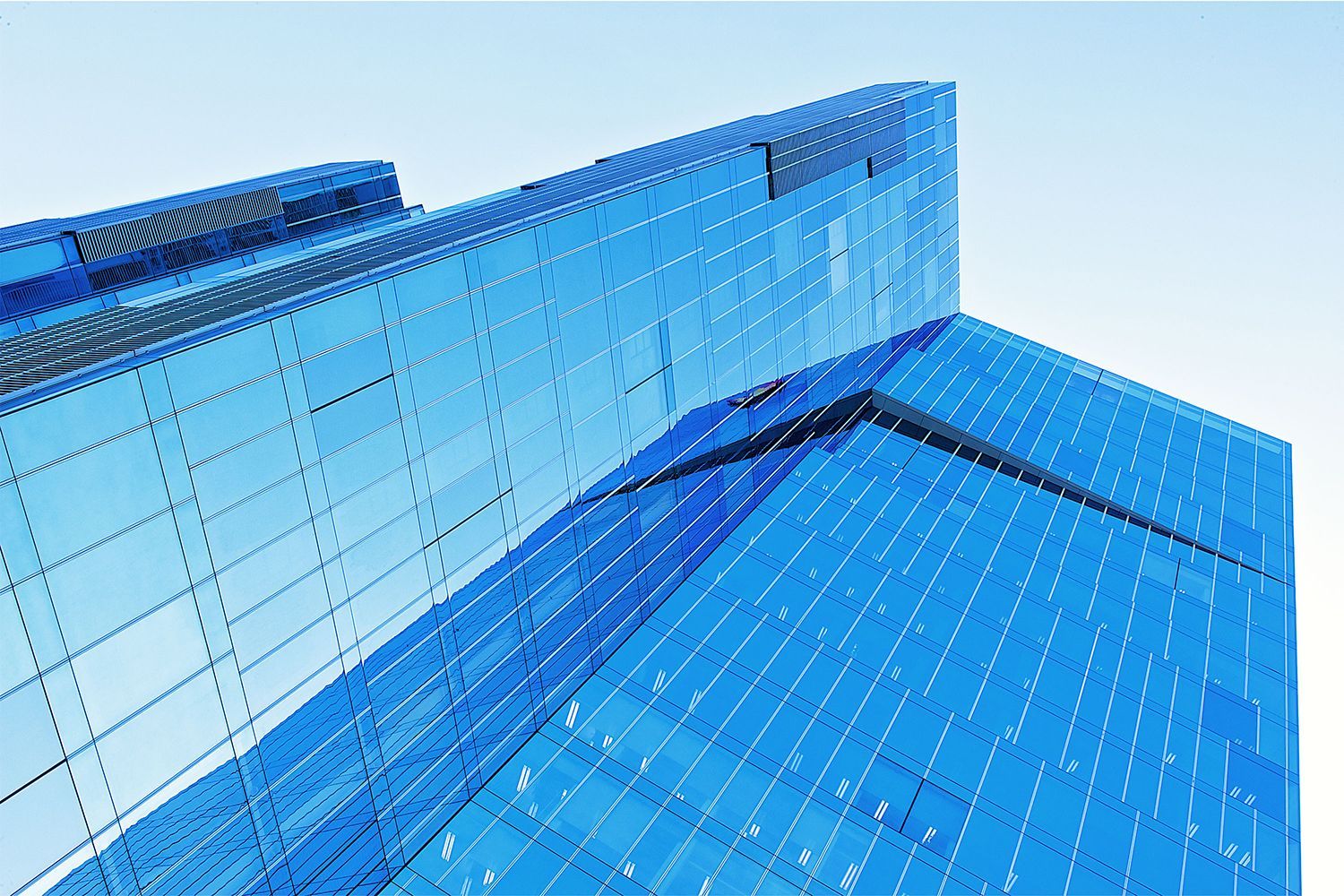 Modern glass skyscraper with a blue hue, angled upwards against a clear sky.