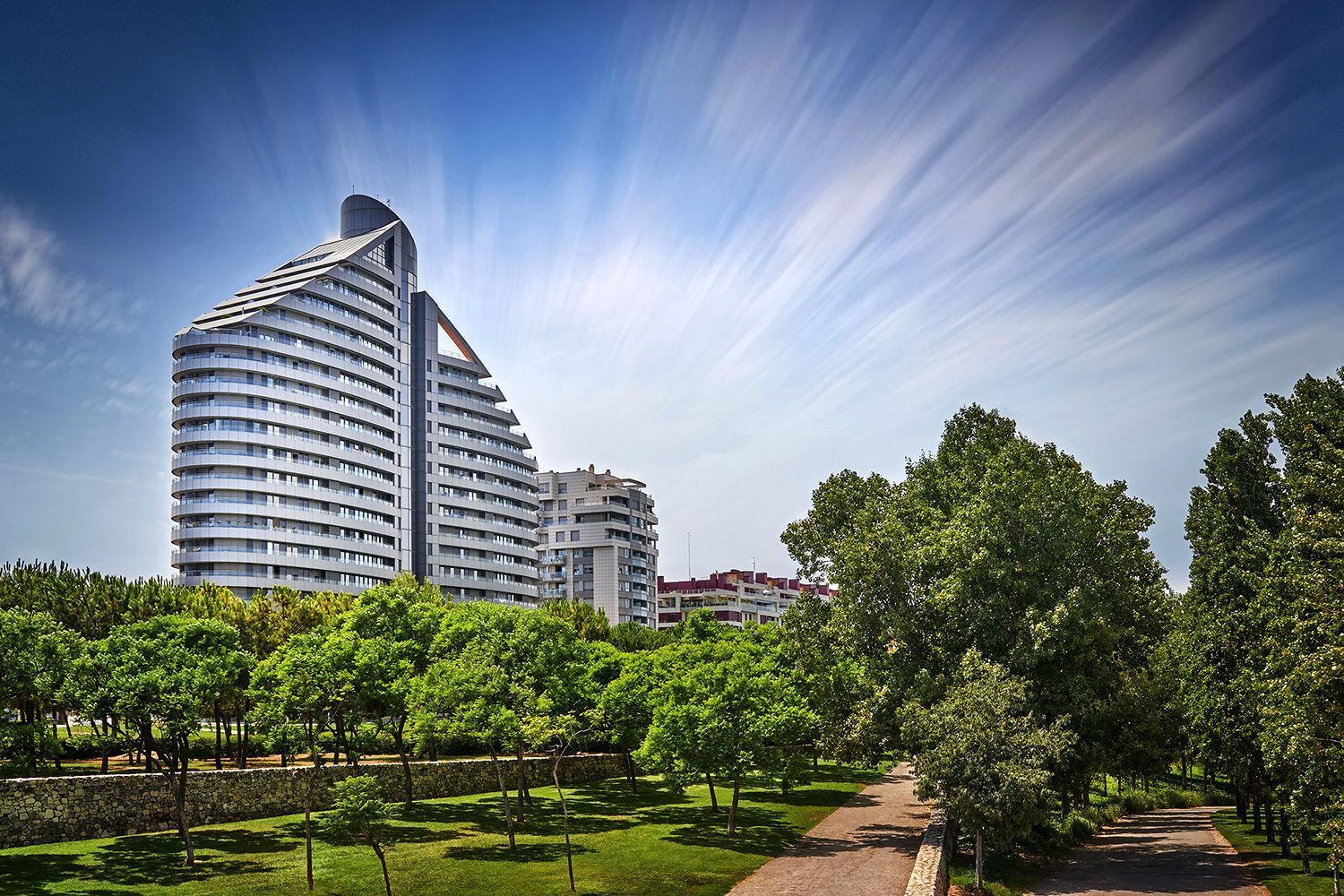 Modern, tiered buildings with white facades rise above a park with green trees and a walking path under a streaked blue sky.