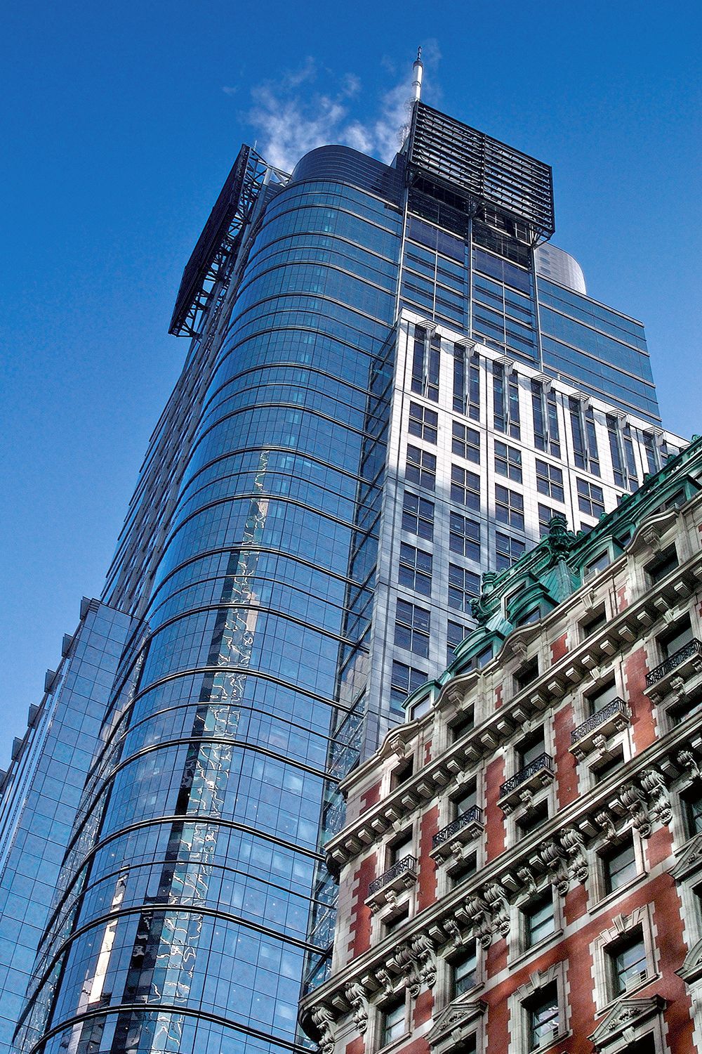 A tall, modern glass skyscraper next to a smaller, ornate, red-brick building under a blue sky.