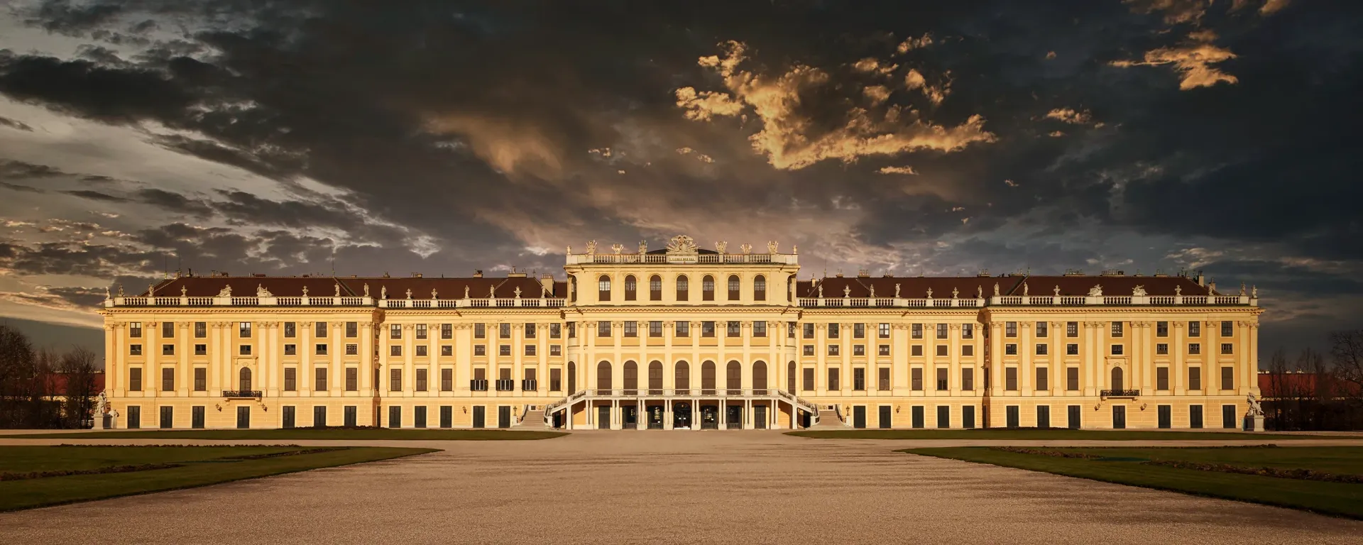 Schönbrunn Palace, Vienna, Austria, at dusk, against dramatic cloudy sky, symmetrical architecture, and open plaza.