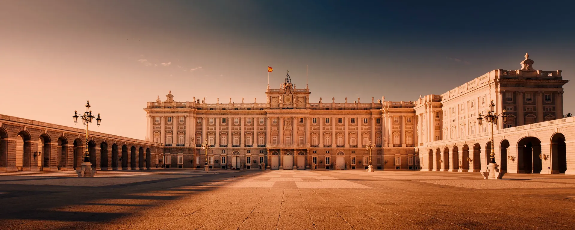 Exterior view of the Royal Palace of Madrid, Spain, with colonnades and a large plaza under a warm, amber sky.