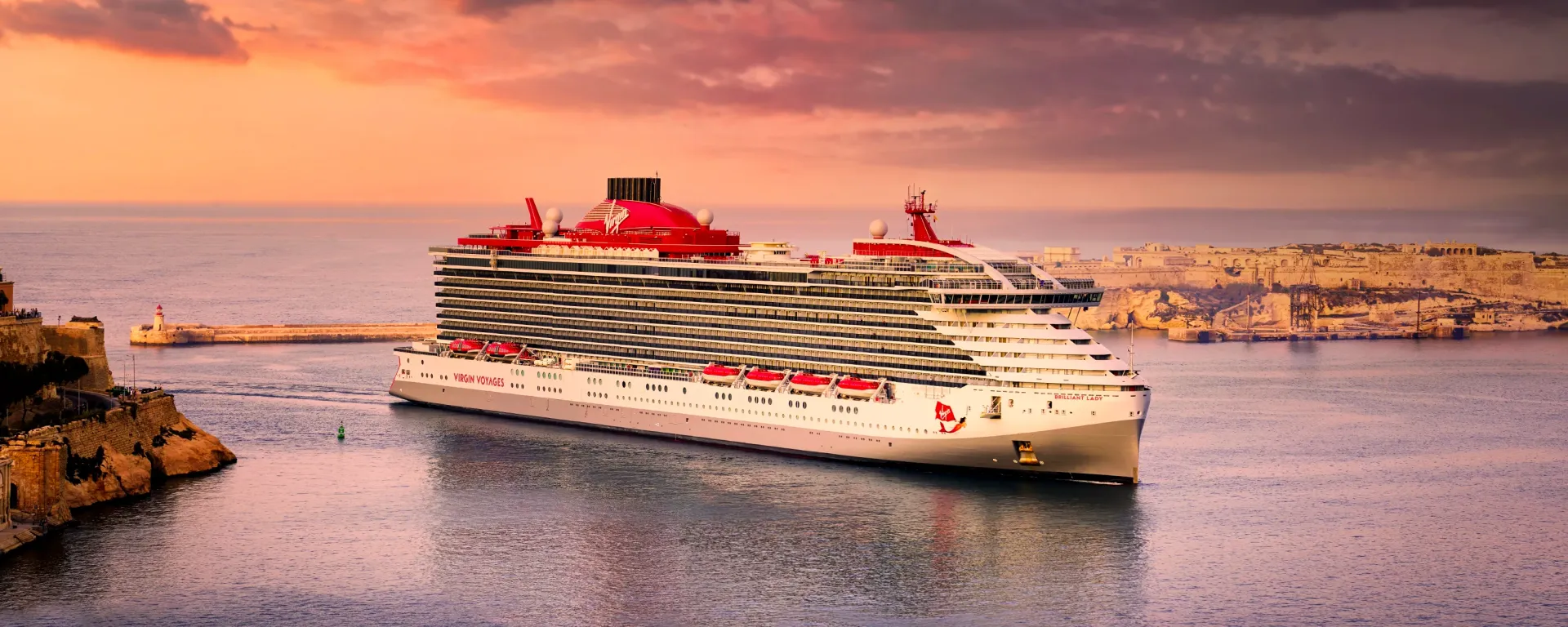 A cruise ship with red accents sails into a harbor at sunset.