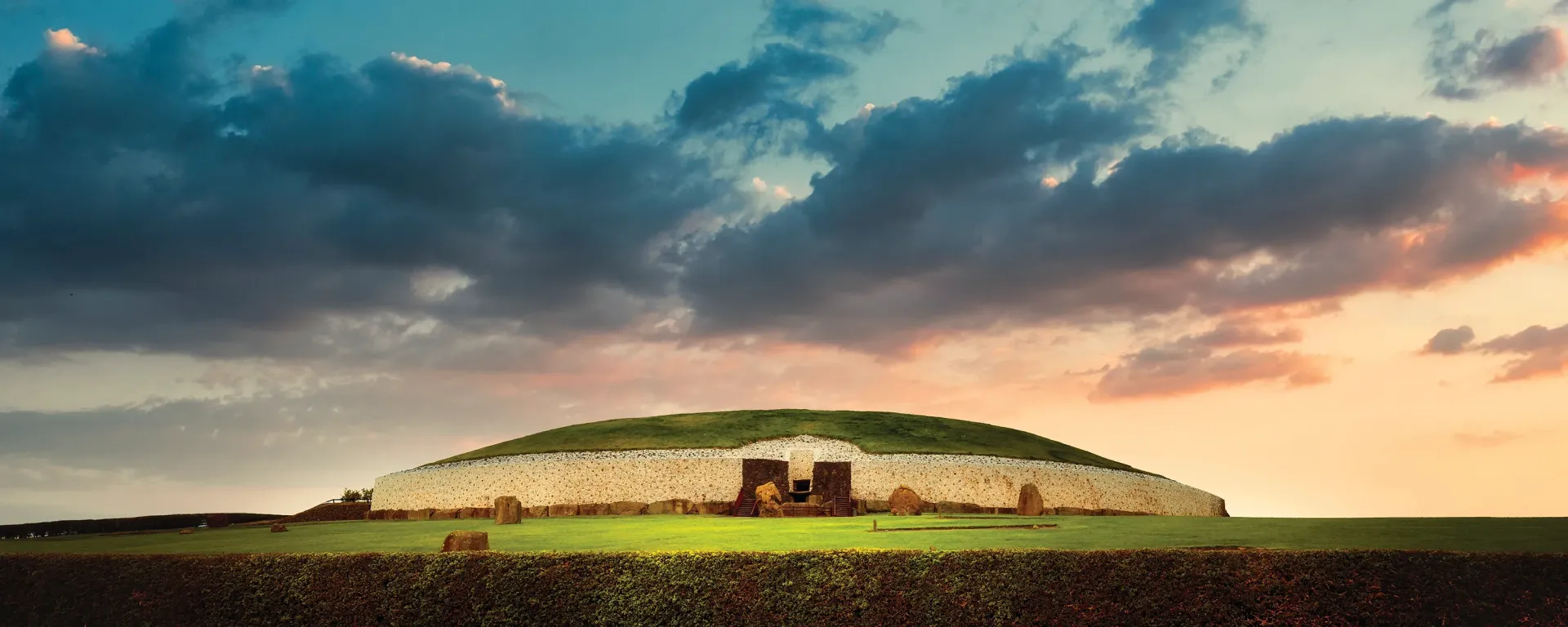 A large, grass-covered mound under a dramatic, colorful sky. Stone entrance visible on the structure's face.