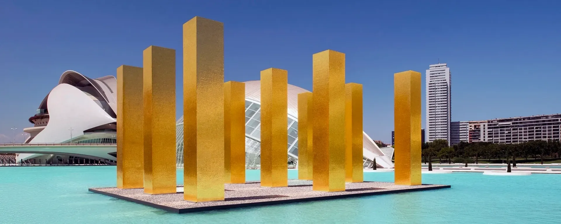 Golden abstract sculpture installation on a floating platform in turquoise water, with modern buildings behind it