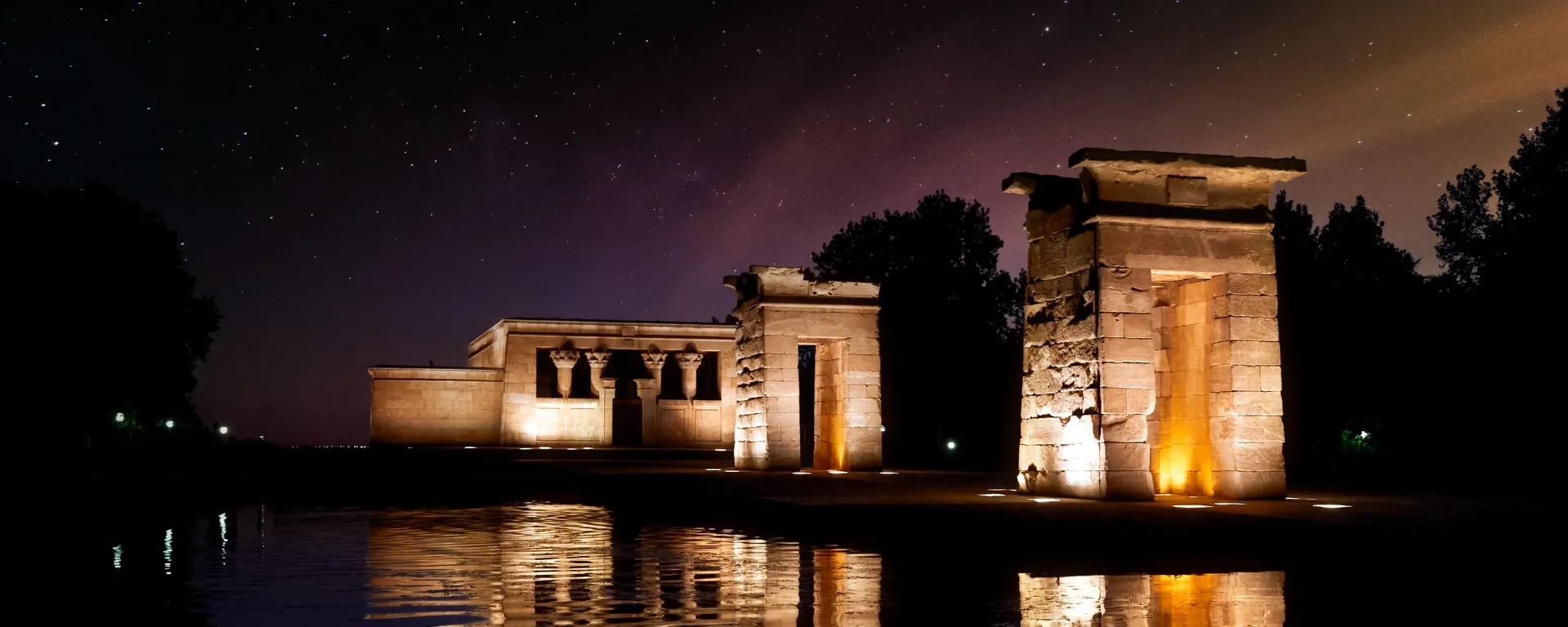 The Temple of Debod illuminated at night, reflecting in the water. Dark sky with stars.